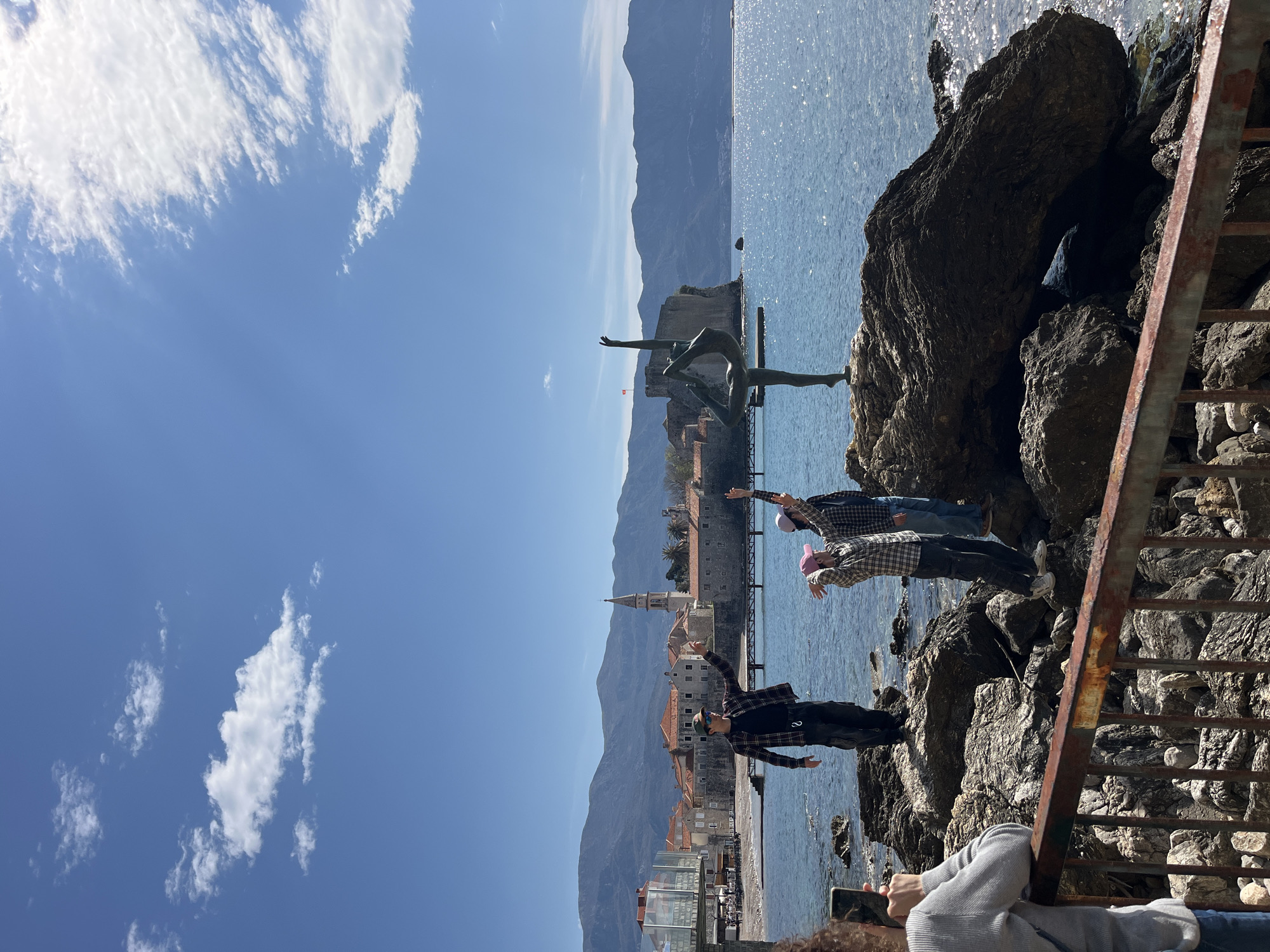 The image shows a coastal scene in Budva, Montenegro. The city is visible in the background, with its buildings and a church spire. A statue stands on a rocky outcrop in the sea. In the foreground, three people are standing on rocks near a rusted metal railing, with their arms raised towards the statue. The sky is blue with scattered clouds, and the sea is a clear blue. The composition includes a mix of natural elements (rocks, sea, sky) and urban elements (buildings, statue, railing).