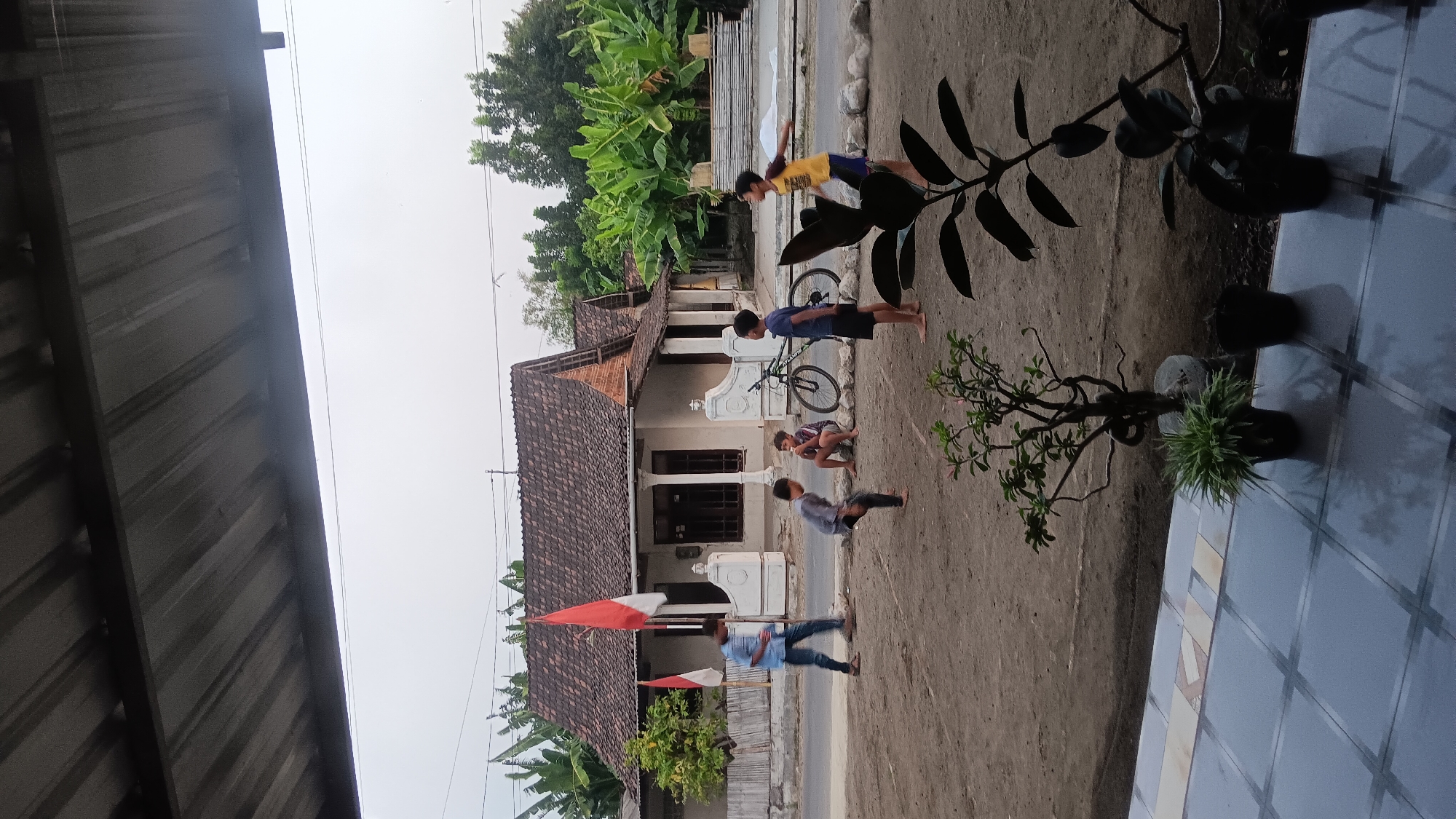 This is a medium shot, eye-level, vertical photograph of a street scene in Sonobekel, Indonesia. The foreground features a tiled patio with several potted plants. The middle ground shows a dirt area where five young boys are playing. One boy in a blue shirt and jeans is walking. Two boys are in motion, one appearing to kick a ball and another with his arms raised. A fourth boy is sitting while eating, and a fifth boy in a yellow shirt is standing. A bicycle is parked on the side of the road. Two Indonesian flags are visible in front of a white house with a brown tile roof. The background consists of trees and a cloudy sky. The overall lighting suggests it is late afternoon or early morning.