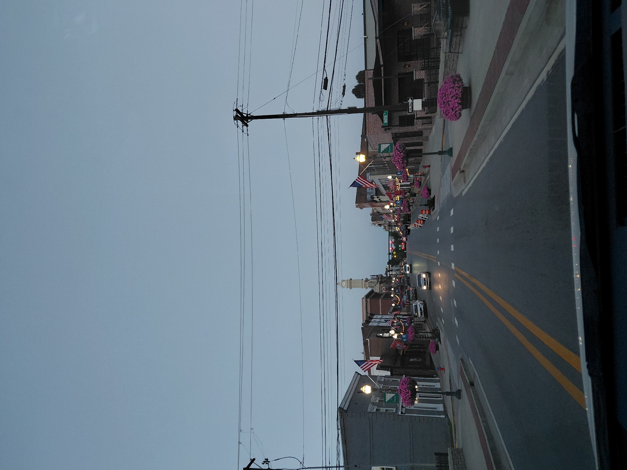 The image depicts a street scene in Georgetown, United States of America, at dusk. The viewpoint is from a vehicle, looking forward down a paved road with double yellow lines. On either side of the road are buildings with architectural styles suggesting a historic downtown area. Many of the buildings are adorned with American flags and decorative banners. Flower baskets filled with pink flowers hang from lampposts and on the sidewalks. Traffic lights are visible in the distance. A tall, ornate clock tower stands prominently in the background, a likely landmark. Several cars are on the road, indicating moderate traffic. The sky is overcast and a pale blue, typical of twilight. Utility poles and overhead wires are visible throughout the scene. The overall impression is a well-maintained, seemingly active, small-town main street.