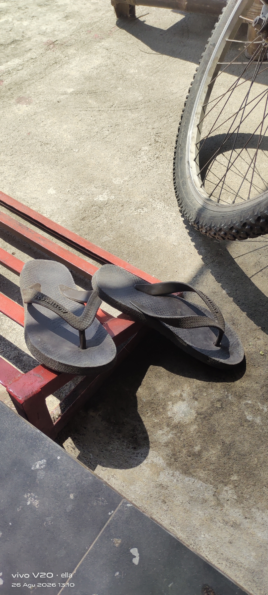 This is a photograph taken outdoors on a concrete surface, likely in the city of Simo, Indonesia, as indicated by the timestamp. The scene is illuminated by bright sunlight, casting distinct shadows. In the foreground, a pair of dark-colored flip-flops are resting on a red metal bench. One flip-flop is positioned with its sole facing upwards, revealing a textured pattern and some branding. The other flip-flop is lying on its side. To the right of the flip-flops, a portion of a bicycle wheel is visible. The tire has a rugged tread, and the spokes are clearly defined. The background consists of a worn concrete ground with some scattered debris. The overall impression is one of a casual, everyday outdoor setting.
