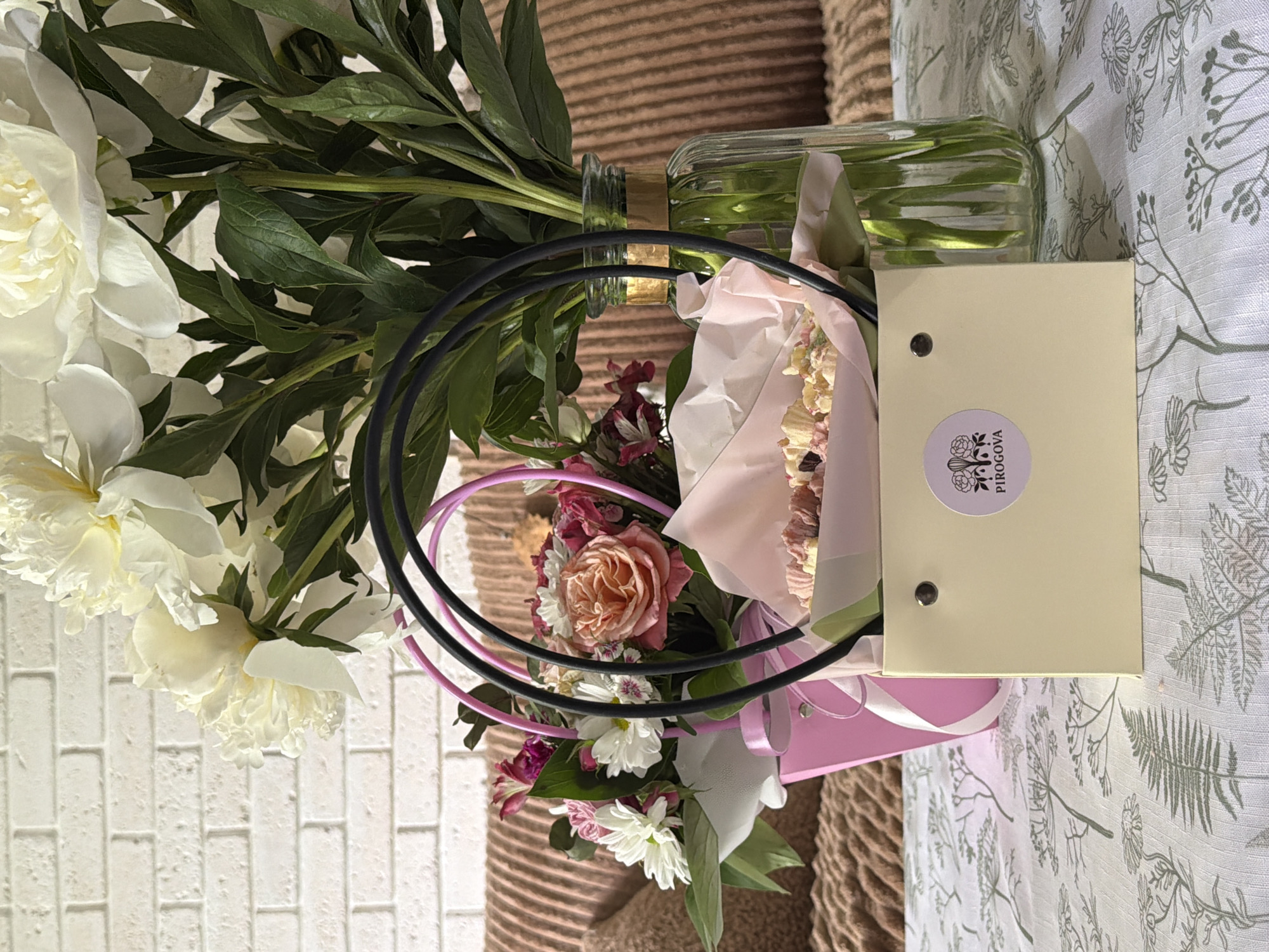 The image shows a floral arrangement with a gift box. The arrangement includes white peonies, a rose, and other smaller white and pink flowers. The flowers are arranged in a clear glass vase and a pink container. A black and pink hoop surrounds the flowers. A light yellow gift box with a round sticker is placed next to the arrangement. The background consists of a white brick wall, a textured brown surface, and a tablecloth with a botanical print. The city and specific location are unknown.