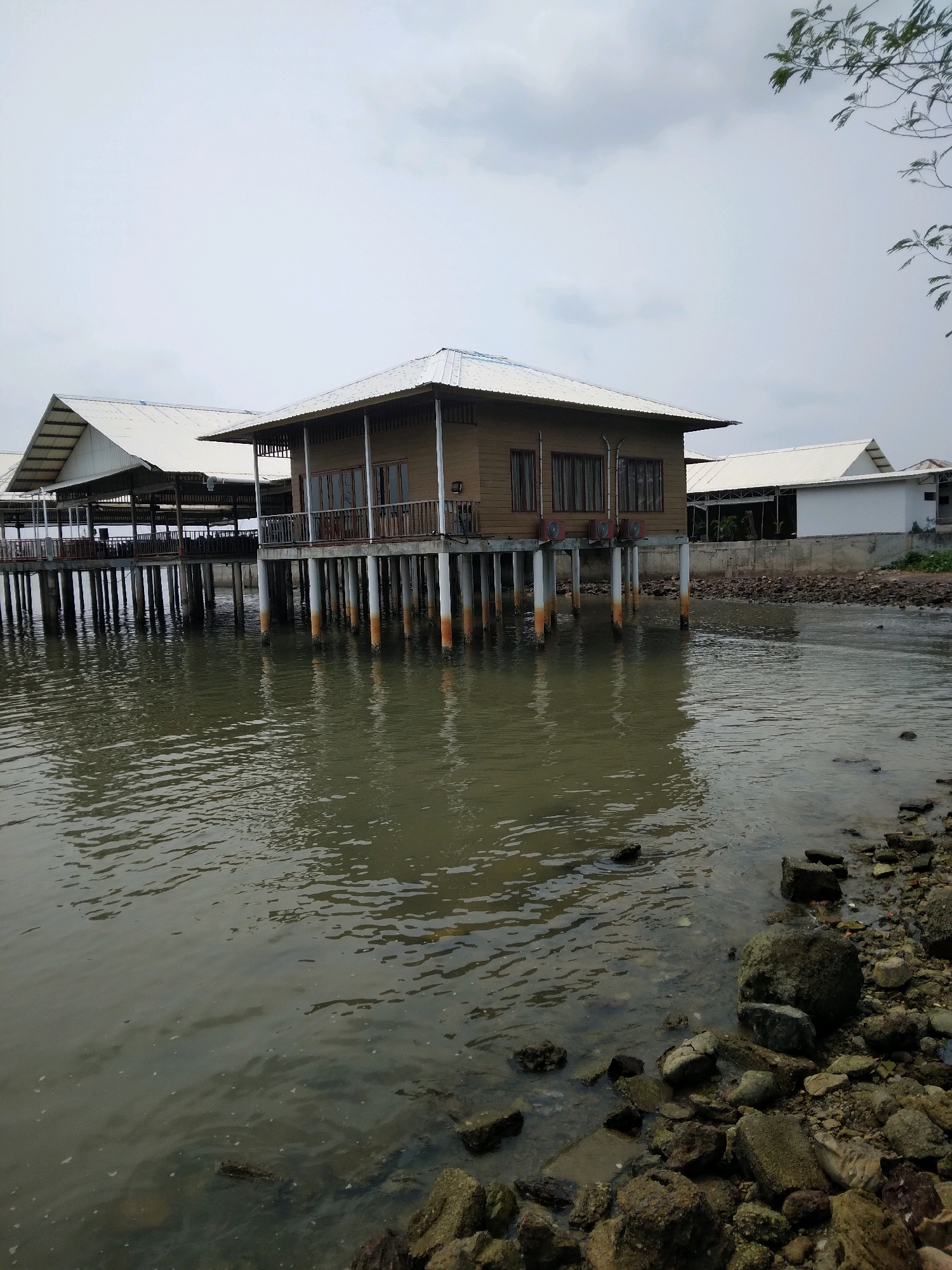 A waterfront view features several stilted buildings and a rocky shore under a cloudy sky. The main building, a light brown wooden structure with a white roof, is supported by white pillars. It has multiple windows with dark frames. To its left, a larger building with a white roof and dark support beams is visible, also on stilts, suggesting a resort or restaurant setting. A white building with a peaked roof stands further back on the land. The water is a murky green, with ripples and a few small whitecaps. The shore is densely covered with various sizes of rocks and some scattered debris. A branch from a tree with green leaves hangs into the upper right corner of the frame. The scene appears to be the waterfront of a coastal area in Batam, Indonesia, likely a commercial or recreational establishment. There are no people or animals visible in the image.