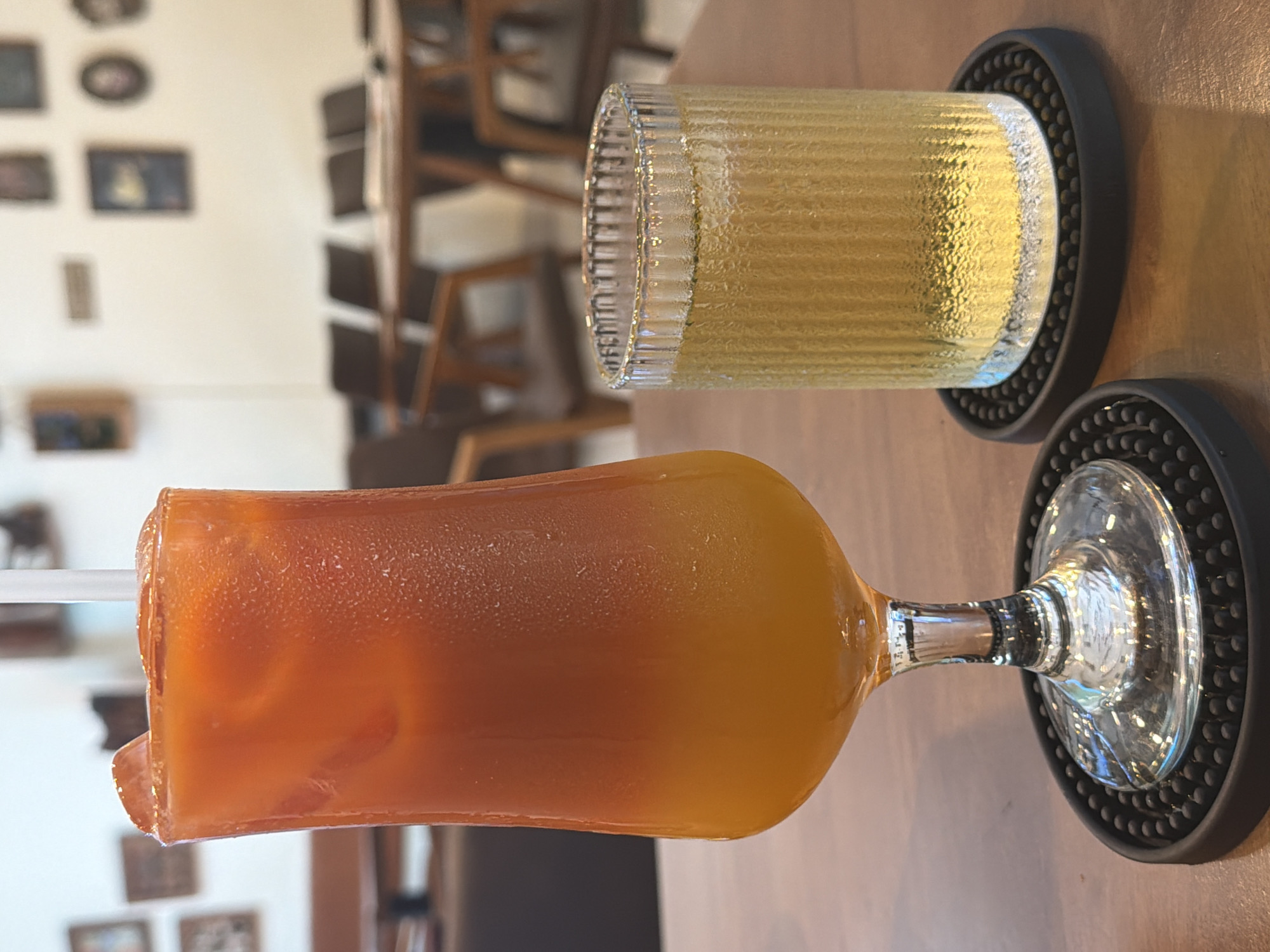Two drinks are on a wooden table in what appears to be a restaurant or cafe setting, possibly in Bac Nha Trang Ward, Vietnam. The drink on the left is in a tall, fluted glass with a wide rim and a thick stem, filled with an orange beverage layered with yellow at the bottom and ice cubes visible at the top, topped with a white straw. The drink on the right is in a shorter, ribbed glass containing a pale yellow liquid. Both glasses are placed on black, circular coasters with a textured surface. In the background, a blurry scene of tables and chairs suggests a dining area, with framed pictures visible on a light-colored wall. The lighting is soft, creating a warm ambiance. There are no people clearly visible in the image.