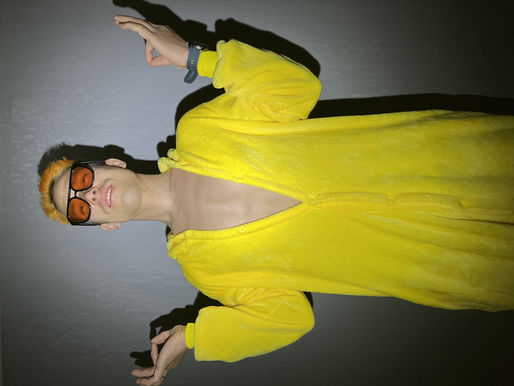 A person wearing a yellow onesie is lying on a grey surface. The person is wearing orange sunglasses and has orange hair. Their onesie is unbuttoned, revealing their chest. A dark blue wristwatch is visible on their left wrist. The location is not discernible.