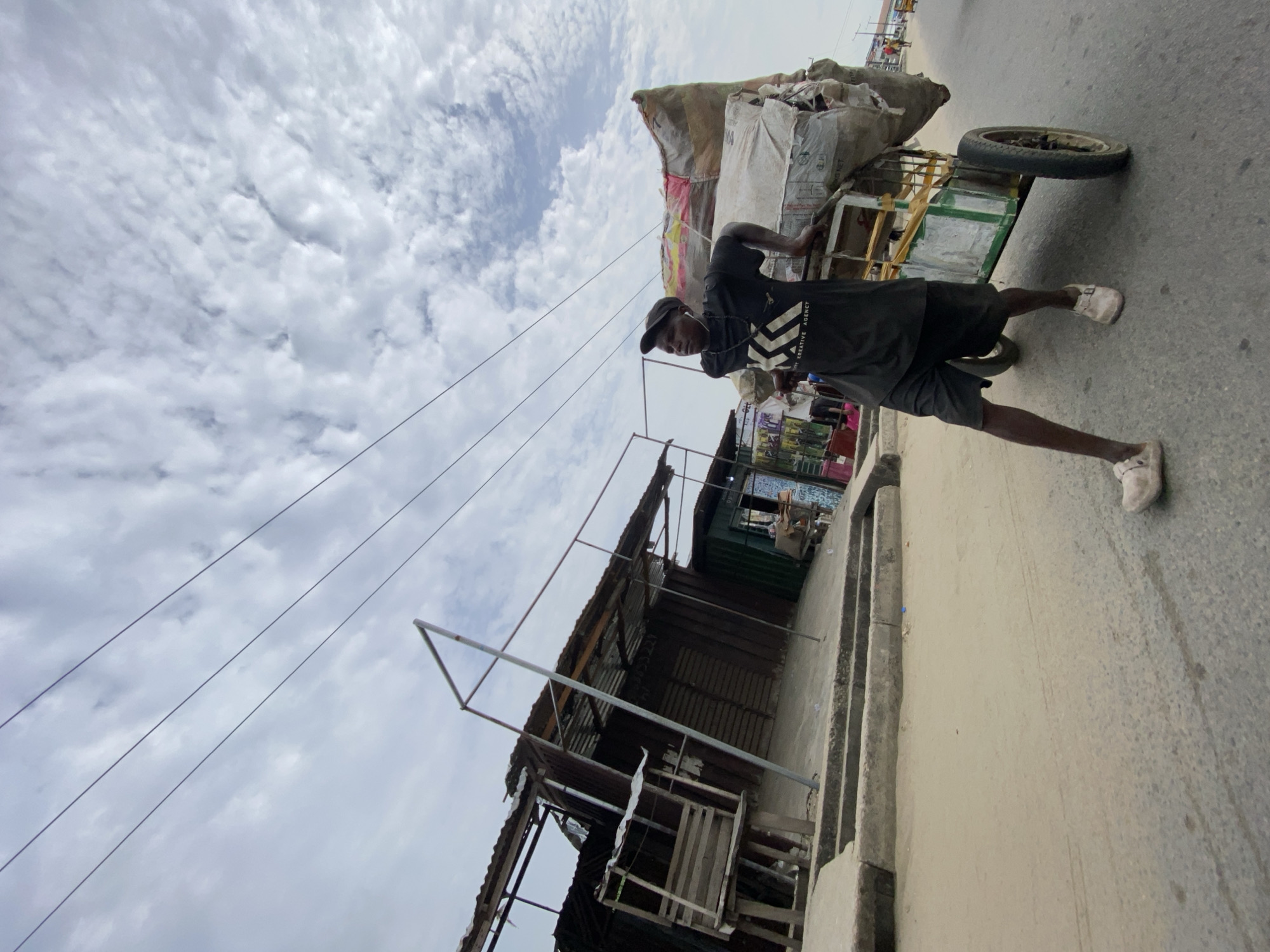 A man in a black shirt, black shorts, a black cap, and white shoes is pulling a green and yellow cart loaded with burlap sacks on a concrete road. To the left of the road are buildings with corrugated metal roofs. The sky is cloudy. The location is identified as Ado, Nigeria, based on contextual information.