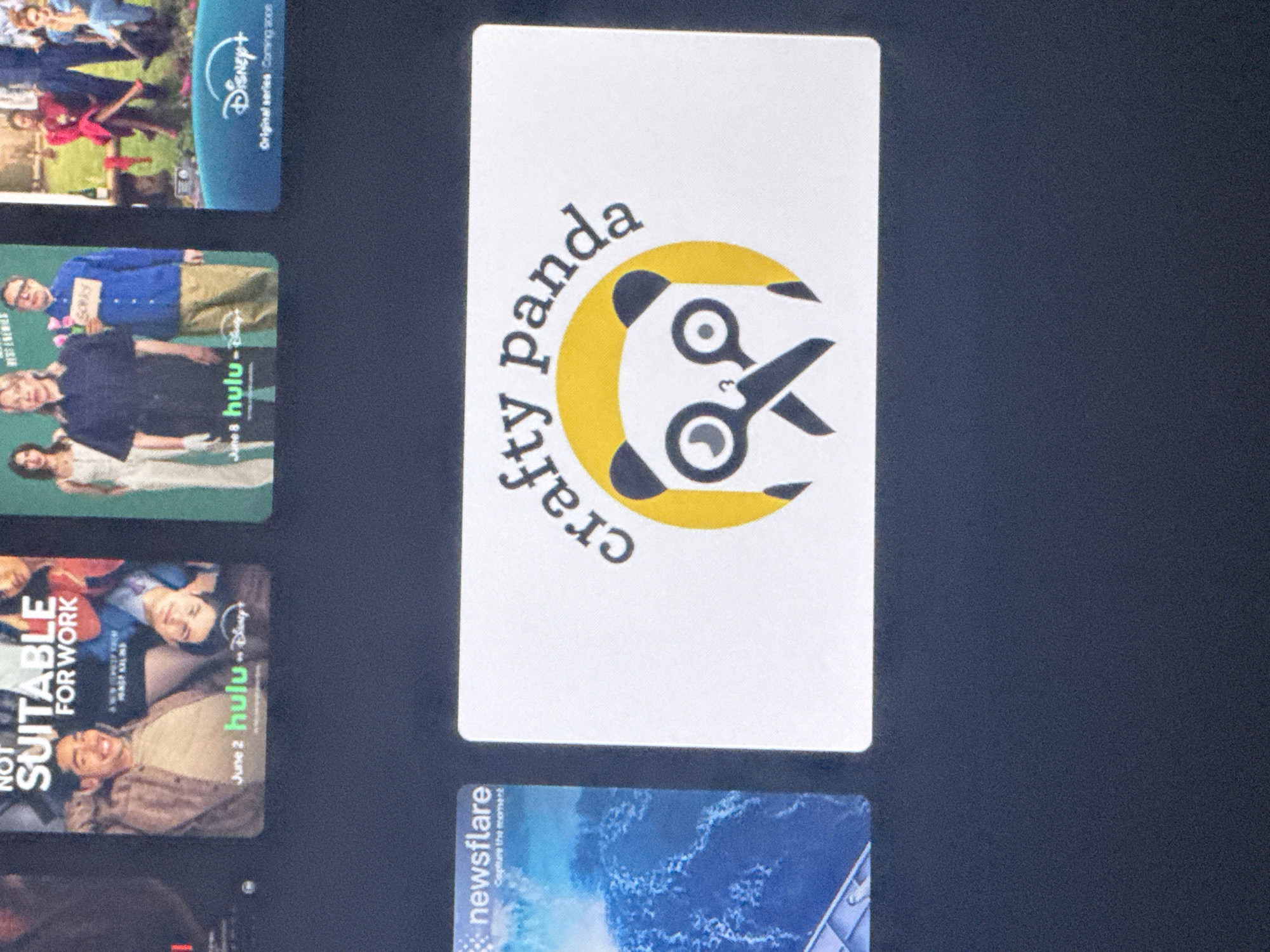 The image shows a close-up, slightly angled view of a television screen displaying various content. In the foreground, a white rectangular card with rounded corners is centered, featuring a logo with the text "crafty panda" arched over a stylized panda face with scissors for eyes. The panda's face is depicted in black and white, set within a yellow circle. To the left of this card, a portion of another card is visible, showing the word "newsflare" and an image of choppy blue water. In the background, across the top of the screen, are sections of what appear to be movie or show posters. One poster is for "NOT SUITABLE FOR WORK," and another features "Disney+ Original series Coming soon." The surrounding environment appears to be a dark room, with the television screen being the primary light source. There is no visible indication of the location being in Saratov, Russia, or any other specific geographic location.