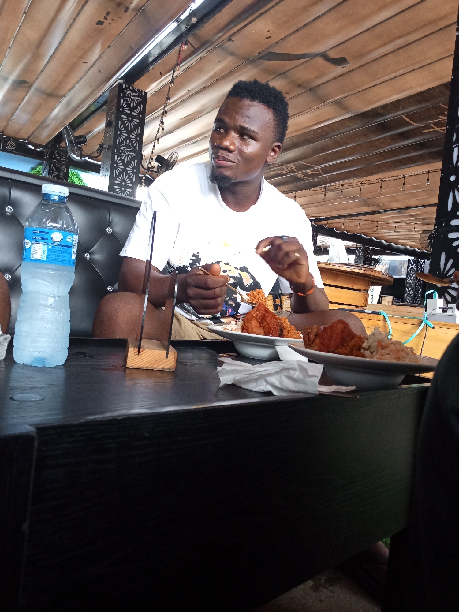 The image captures an indoor or semi-outdoor dining scene within an establishment in Howayi, Nigeria, identified as "Food House" through branding visible on decorative pillars.
In the foreground, a dark, textured rectangular table occupies the lower portion of the frame. On its left, a transparent plastic water bottle with a blue cap and label, marked "STORAGE" and "EAU NATURELLE," stands upright. To the right, two white plates are positioned, each containing a serving of fried chicken (appearing as drumsticks or thighs) accompanied by a portion of orange-colored rice and white coleslaw. Between the water bottle and the plates, a small wooden block serves as a base for two slender, dark vertical slats. White paper napkins are also scattered on the table surface.
An adult male with dark skin, short dark hair, and a short beard is seated behind the table on a black, button-tufted bench. He is wearing a white short-sleeved t-shirt with a dark graphic design and light-colored shorts. He is actively eating, holding a piece of food in his right hand, preparing to bring it to his mouth, while his left hand holds a fork. His head is turned slightly to his right, and his gaze is directed towards the upper right of the frame. A dark-skinned leg and arm, likely belonging to another individual, are partially visible on the far right edge of the frame, indicating the presence of another diner.
The background shows a casual dining environment. The ceiling is constructed from light-colored, horizontal slatted panels. Black decorative pillars featuring white cut-out patterns (floral or star-like) and "Food House" branding are integrated into the structure. A string of small, round light bulbs hangs across the overhead space. A black ceiling fan is partially visible in the upper right corner. Through an opening or window in the far background, green foliage is discernible, suggesting an open-air aspect or view.