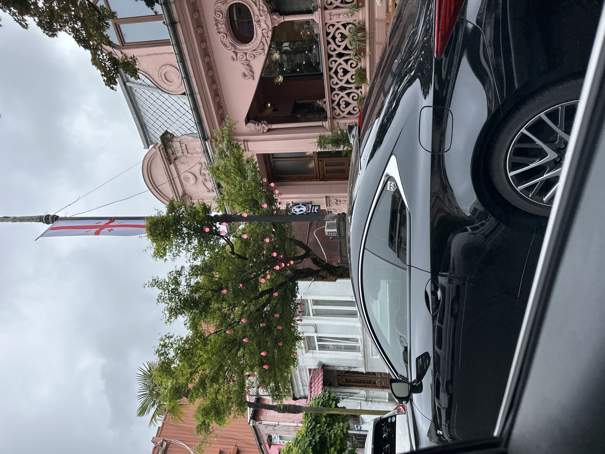 A street scene in Batumi, Georgia, is depicted from the vantage point of inside a vehicle. In the foreground, the side and rear of a black car are visible, with a focus on its bodywork and wheel. Outside the car, a mix of buildings lines the street, including a distinctive pink building with ornate architectural details. A Georgian flag is prominently displayed on a pole alongside a tree adorned with small, pink decorative lights. The sky overhead is overcast, suggesting a cloudy day.
