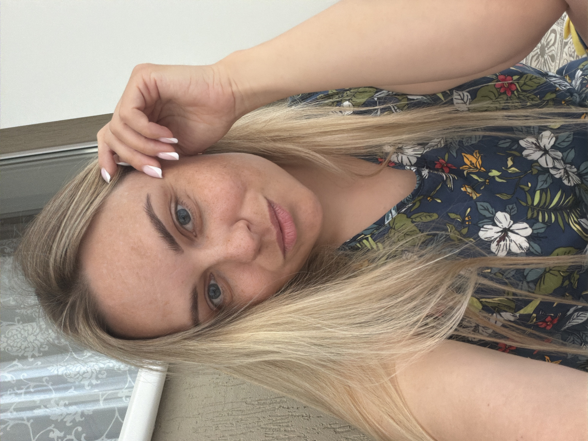 A fair-skinned woman with blonde hair is pictured in a medium close-up. Her hair is long, straight, and falls across her shoulder and part of her face. She has light eyes, freckles, and is looking directly at the camera with a slight smile. Her right hand is raised near her forehead with manicured nails featuring white tips. She is wearing a dark blue floral print top. The background is indistinct but appears to include a window with a patterned curtain. It is not possible to verify that this photo was taken in Votkinsk, Russia.