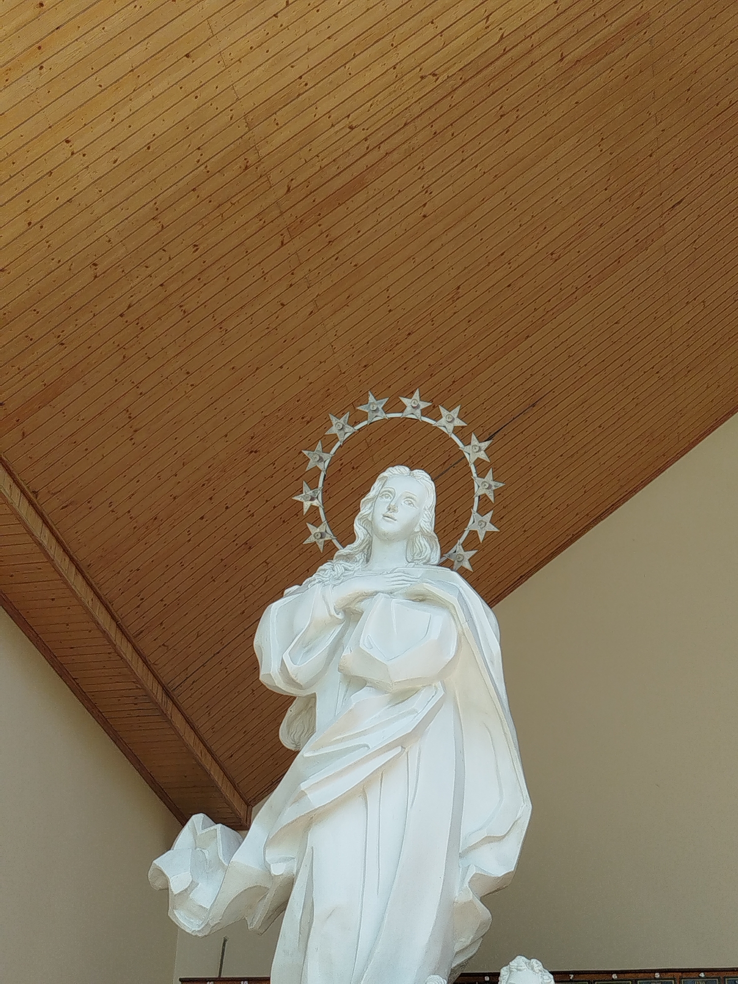 A low-angle shot shows a white statue of a woman with long hair and a star-studded halo against a backdrop of a brown wooden ceiling. The statue depicts the woman in flowing robes, with her hands crossed over her chest. The building is located in the city of Pleiku Ward, Vietnam.