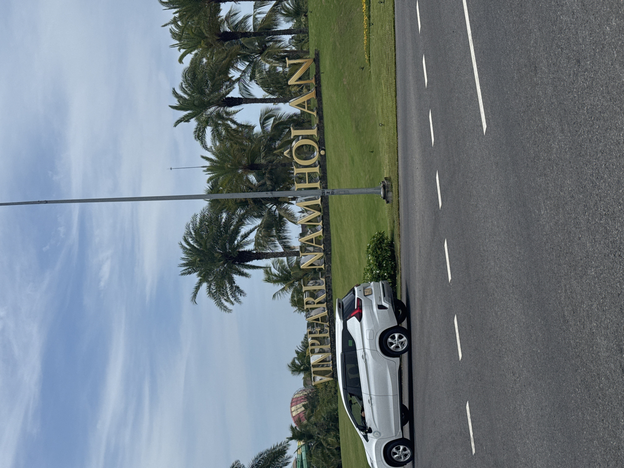 A white Mitsubishi Xpander is parked on the side of a road in Đà Nẵng, Vietnam. The car is next to a grassy area with palm trees and a large sign that reads "VINPEARL NAM HOI AN." The sky is blue with some clouds. The road has white dashed lines.