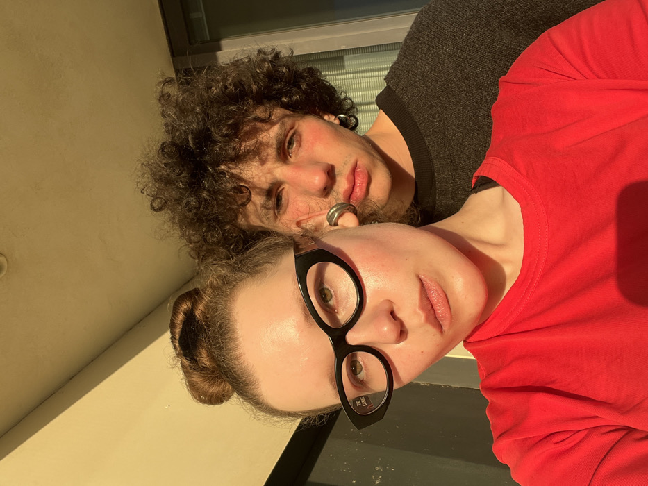 The image presents two individuals, a woman and a man, captured in a close-up, front-facing composition under strong, warm directional sunlight. The setting appears to be an urban exterior, likely a balcony or building facade in Tel-Aviv, Israel, though specific geographical markers for a precise location within the city are not visible.
In the foreground, a woman with light skin and reddish-brown hair styled in a high bun wears black, cat-eye eyeglasses, on which "THE CLIQUE" is printed on the left lens. Her eyes are open and directed towards the camera, and her lips are slightly parted. She is dressed in a bright red crew-neck t-shirt. A silver cuff-style earring is visible on her left earlobe.
Positioned behind and slightly to the right of the woman is a man with light skin, dark, voluminous curly hair, and visible redness on his cheeks and the bridge of his nose. He wears a dark grey crew-neck sweater. His eyes are open, also looking directly at the camera, and his lips are closed with a neutral expression. Two silver hoop earrings are visible in his left ear.
The background consists of a plain, light-colored (beige/cream) wall or ceiling structure in the upper left, transitioning to a vertical element on the right side behind the man. This vertical element displays horizontal striations, suggesting a window or door with blinds or slats. The lighting indicates direct, low-angle sunlight, consistent with either early morning or late afternoon, casting warm, golden tones across the subjects.