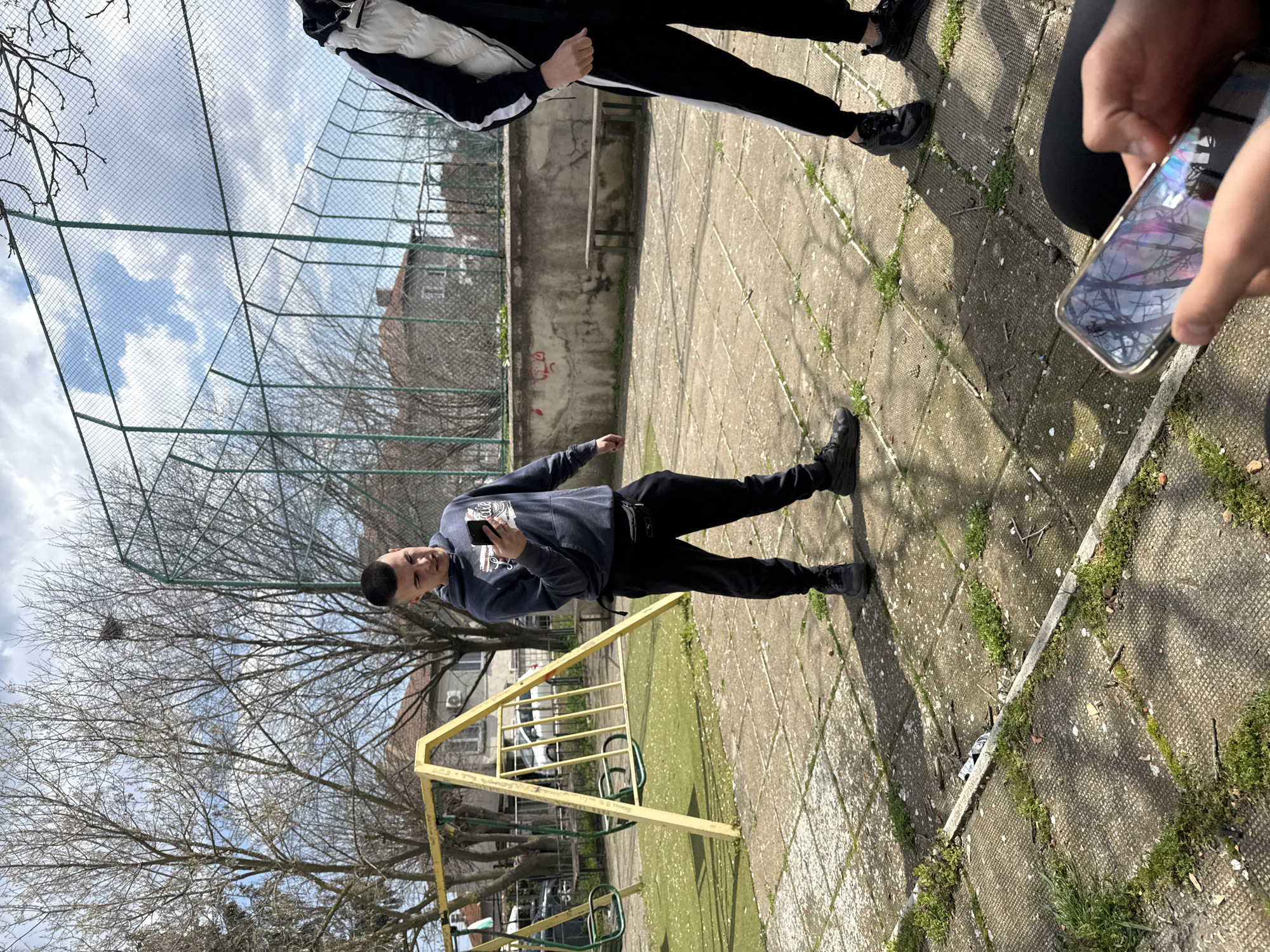 The image shows a playground area, likely in Topoli, Bulgaria. A young man is standing on a paved surface, holding a smartphone and appearing to take a picture or video. He is wearing a dark blue hooded sweatshirt and dark pants. Other individuals are partially visible in the periphery, including a person with dark pants and shoes and someone holding a smartphone. A yellow swing set is present. The background includes a chain-link fence, buildings, and bare trees suggesting a late fall or winter season. The pavement has weeds growing in the cracks.