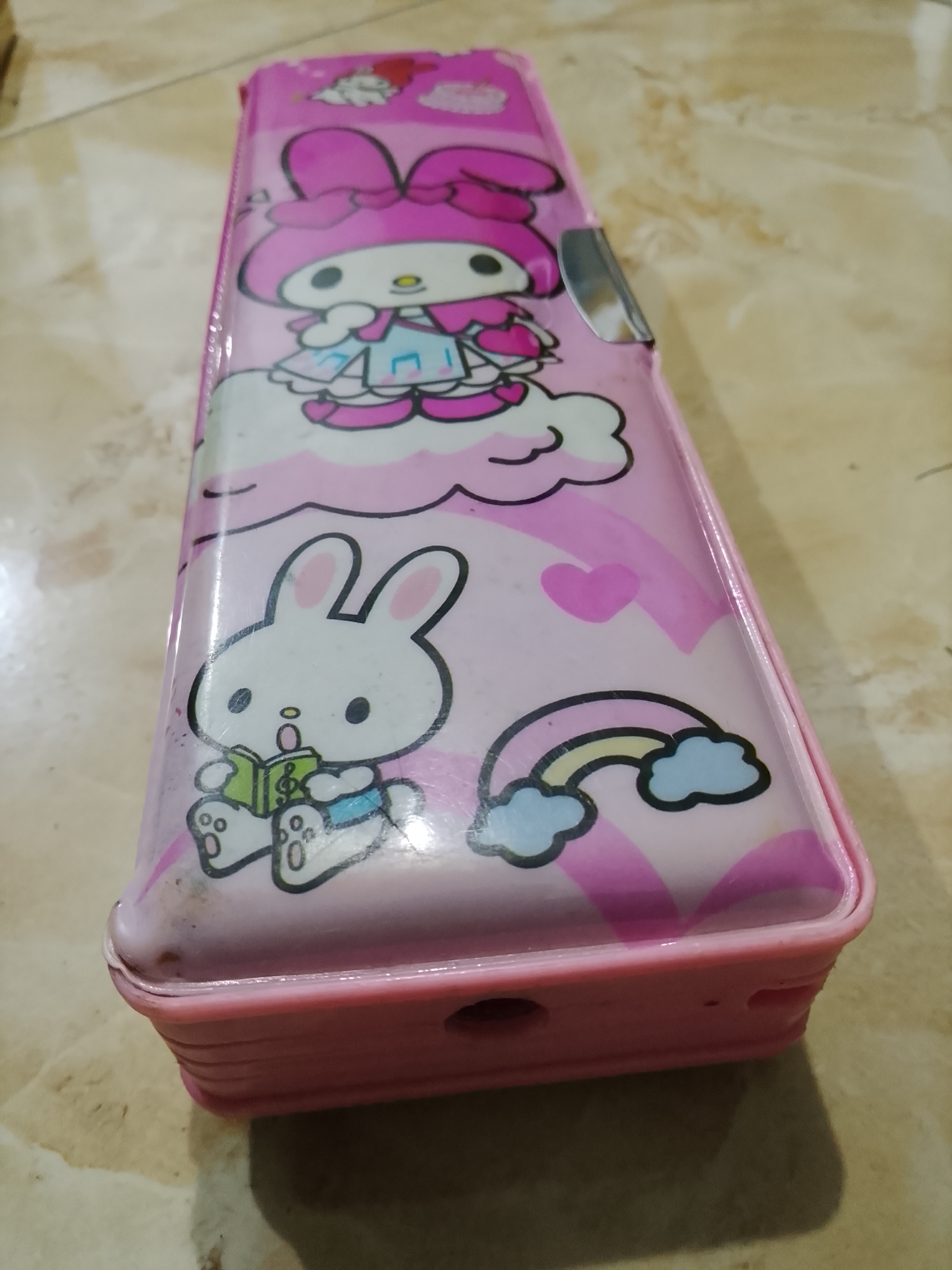A pink pencil case with cartoon characters is displayed on a beige tiled surface. The pencil case features illustrations of a bunny reading a book and a character resembling My Melody. The pencil case is rectangular with rounded edges and has a silver clasp on its side. The tiled surface appears to be a floor. The image was taken in Dawuan, Indonesia.
