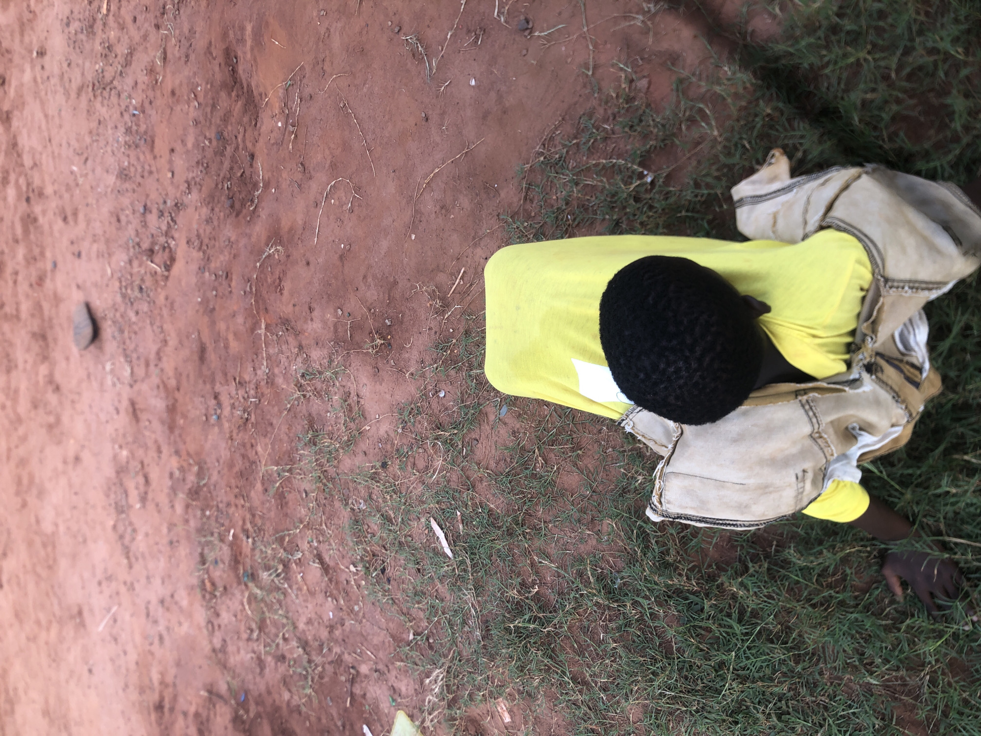 An overhead photograph captures a person sitting or kneeling on reddish-brown earth interspersed with patches of green grass, likely in an outdoor setting in Nsugbe, Nigeria. The individual is viewed from behind, their head covered in short, dark, tightly curled hair. They are dressed in a vibrant yellow top, layered with a lighter, possibly faded denim or canvas garment, detailed with visible seams and stitching. One dark-skinned hand is visible resting on the grass in the lower right corner of the frame.
The ground features exposed red soil, dotted with small pebbles, scattered dry grass, and healthy clumps of green grass. A single dark grey stone lies on the bare earth in the upper left. The lighting indicates it is daytime. The person appears stationary, suggesting they might be resting or engaged in a quiet activity. No specific emotions are discernible from this vantage point, and no text is visible in the image.