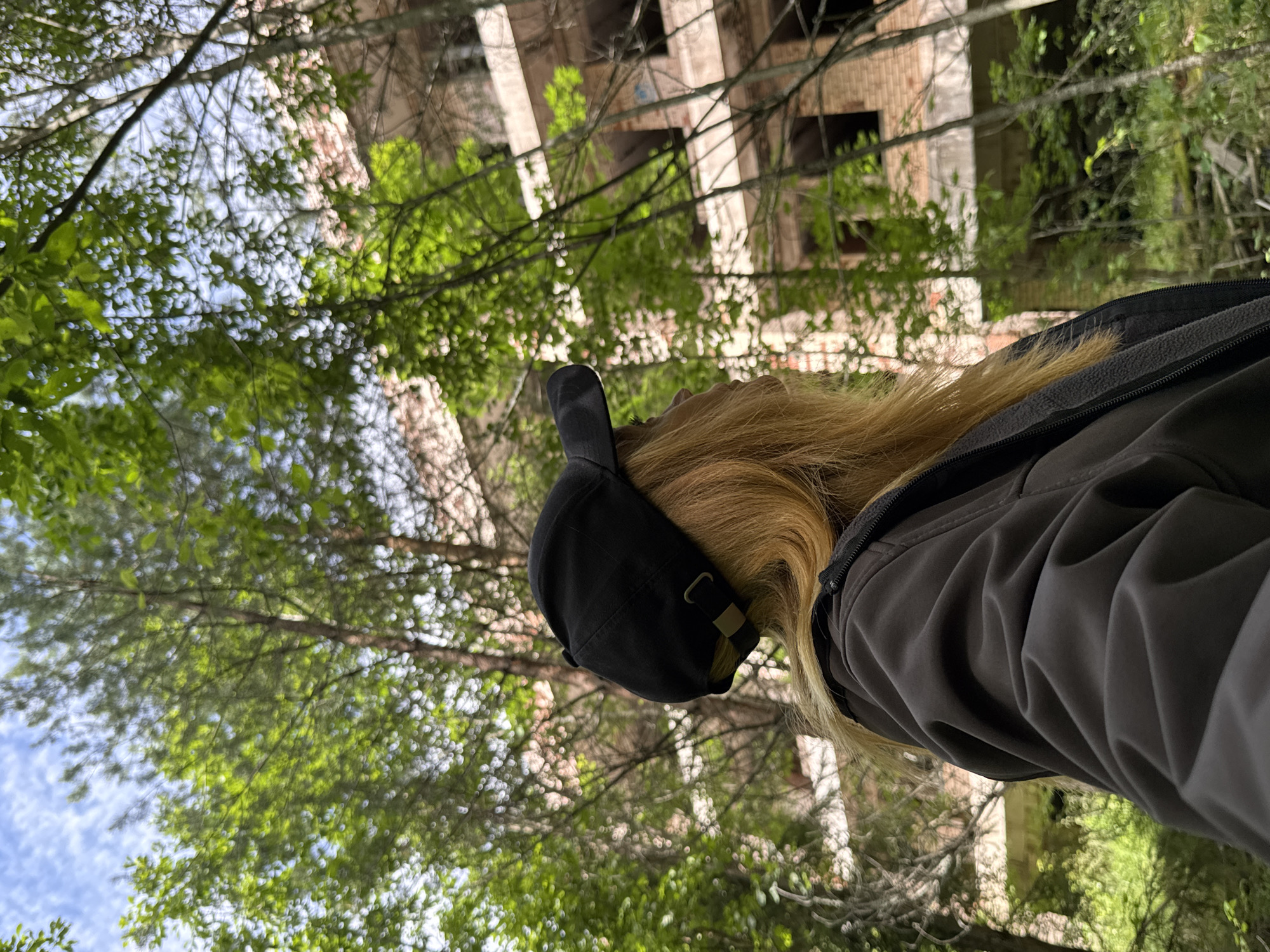 In the city of Interkompleks, Russia, a person wearing a black baseball cap and a dark jacket is viewed from behind, looking towards an abandoned brick building overgrown with trees. The building appears to be several stories high with visible windows and balconies. Lush green foliage surrounds the structure, with sunlight filtering through the leaves, creating a dappled effect on the scene. The sky above is partly cloudy, with patches of blue visible. The overall impression is one of decay and nature reclaiming an urban environment.