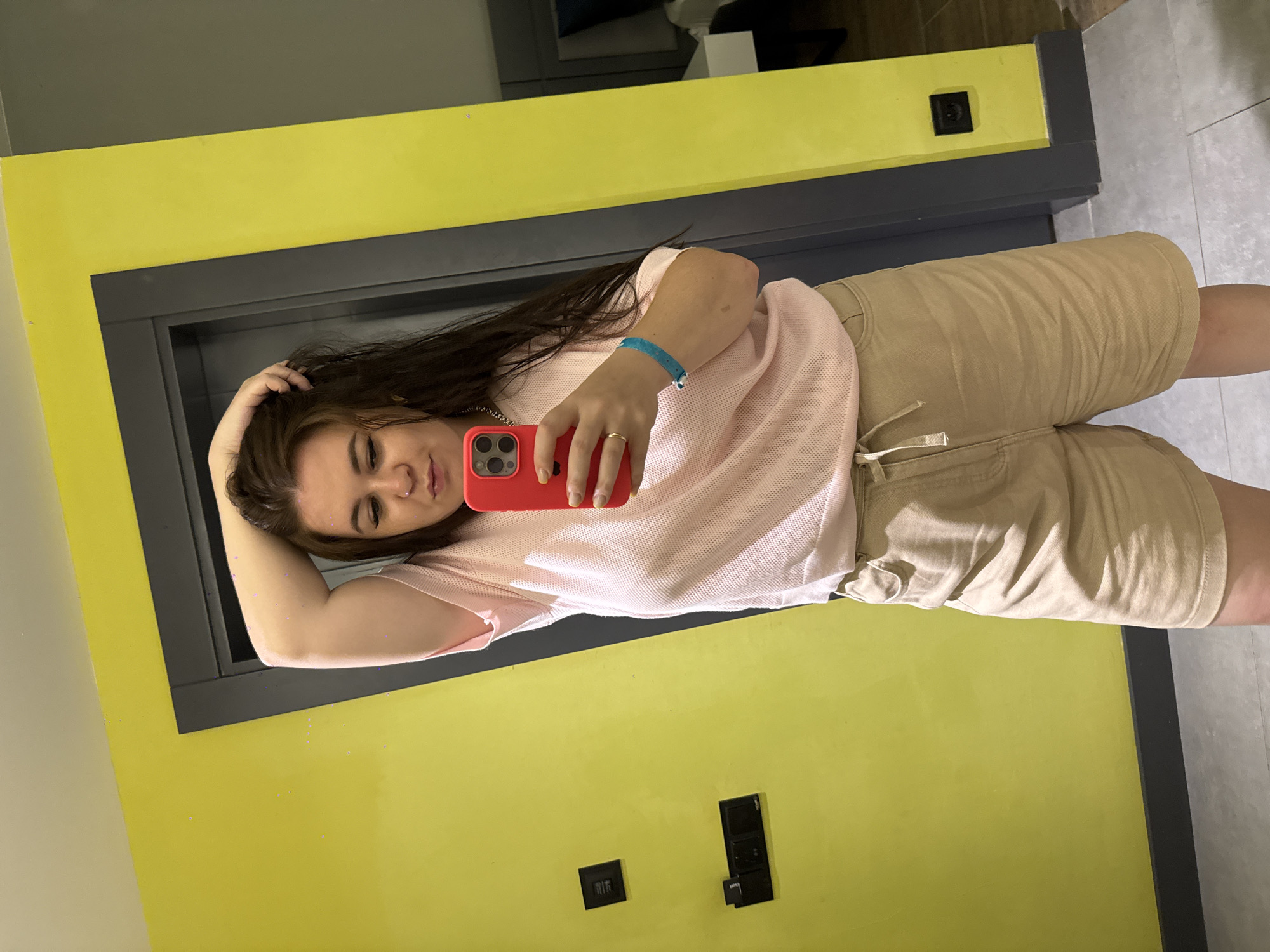 A young woman is taking a mirror selfie in a room with bright yellow walls. She is wearing a light pink mesh-like t-shirt and light brown shorts. Her right arm is raised, and she is touching her hair. She is holding a red smartphone in her left hand. She has a blue bracelet on her left wrist. A gold ring is visible on her left ring finger. The room appears to be a hotel room, with a doorway visible behind her. The floor is tiled. There are two black electrical outlets on the yellow wall to her left. The overall impression is a casual, personal moment captured in a temporary living space. There is no specific indication in the image to suggest the location is Alanya, Turkey.