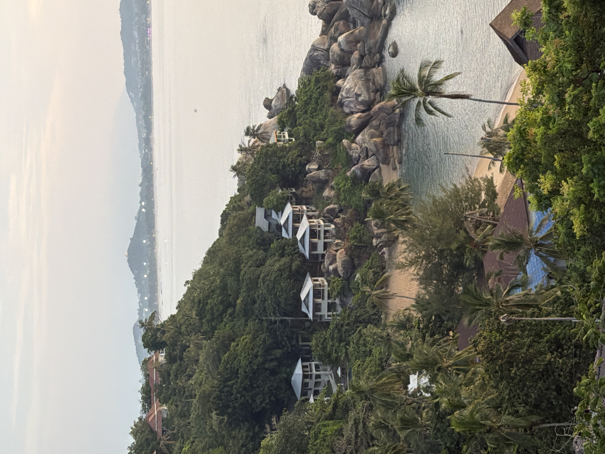 This is a high-angle, full shot of a tropical coastline in Ko Samui District, Thailand. The scene is dominated by lush green vegetation, including numerous palm trees, that cover a steep hillside leading down to a rocky beach and the ocean. Several multi-story villas with white exteriors and grey roofs are nestled into the hillside, overlooking the water. A sandy beach is visible between the villas and the sea, with a single palm tree standing tall on the sand. The ocean is calm and reflects the hazy, pale sky, which suggests either early morning or late afternoon light. In the distance, more hills and coastline are visible, with some buildings and lights indicating a settlement. A small boat is present on the water.