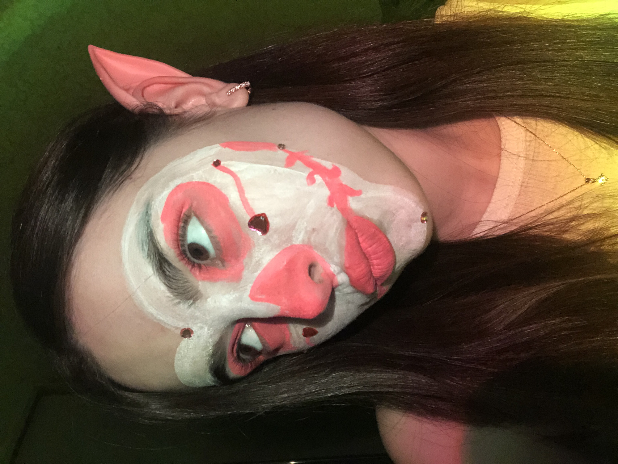 A person with pig-like makeup and an attached pig ear is lying down. The makeup consists of white base paint, pink accents for the nose and around the eyes and mouth, and small red gemstone-like embellishments. The person has long, dark hair and is wearing a light-colored top. A gold necklace is visible on the person's neck. The background appears to be a dark green hue. The location is not evident from the image.