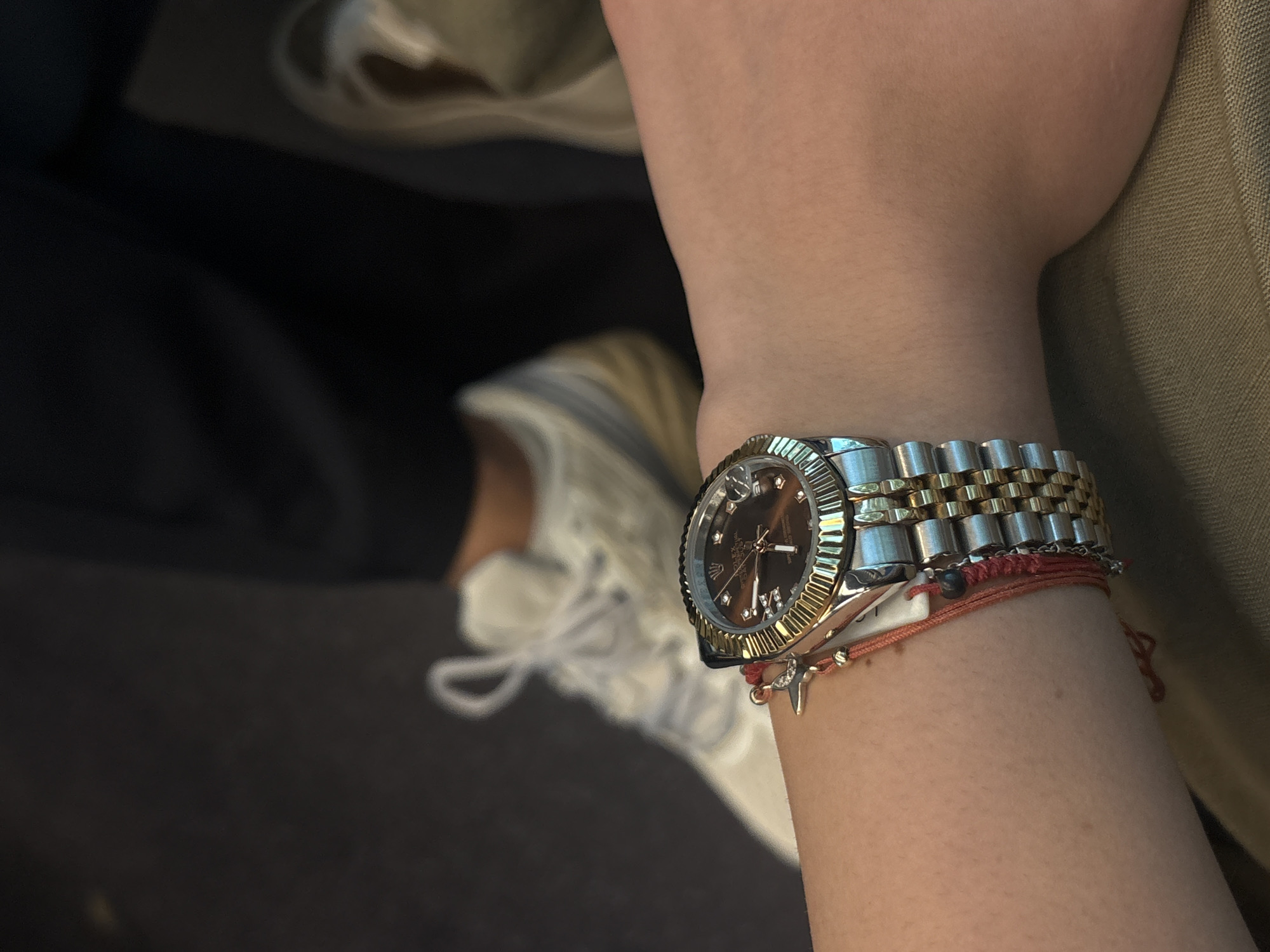 The image shows a person's wrist wearing a two-tone Rolex Datejust watch with a brown dial and fluted bezel. The watch band is two-tone, silver and gold. Adjacent to the watch are two thin, braided bracelets in shades of red and orange, adorned with small charms. In the background, there is a beige sneaker with white laces and a portion of dark fabric, possibly part of clothing. The setting appears to be indoors, likely a casual or personal space. There is no information to suggest that the image was taken in Alessandria, Italy.