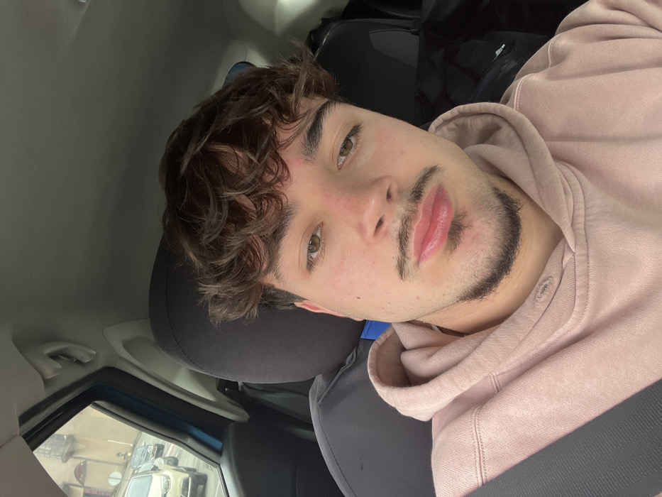 A male subject is depicted in a close-up, front-facing image, seated within the driver's compartment of a vehicle. He exhibits medium-length, dark brown, curly hair, light brown eyes, and fair skin with observable erythema on the forehead and cheeks. Distinct facial hair includes a thin mustache extending to a chin strip. The subject's lips are full. He is attired in a light pink hooded sweatshirt and a dark-colored seatbelt diagonally across his torso. The vehicle's interior shows a dark headrest and a light blue A-pillar. Through the front left window, an exterior scene is visible, consisting of a paved roadway or parking lot with multiple stationary vehicles and a multi-story, light-colored building featuring a balcony. This location is identified as Santa Marta de Penaguião, Portugal, under ambient lighting conditions suggesting overcast weather.