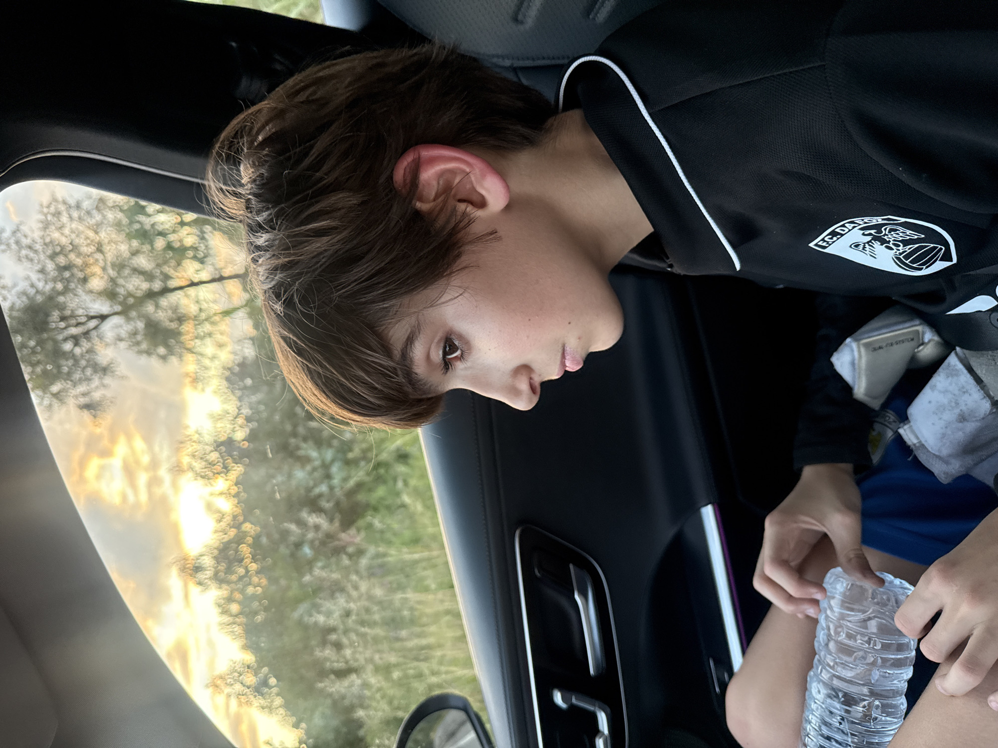 A youth is seated in a vehicle. The youth has dark hair, fair skin, and is wearing a black soccer jersey with white trim and the FC Dakota logo. The youth holds a plastic water bottle on their lap. Outside the car window is a blurred scene of vegetation with a sunset. The vehicle's door and mirror are visible. The location is unknown and cannot be determined to be in Vila Nova de Gaia, Portugal, from this image alone.