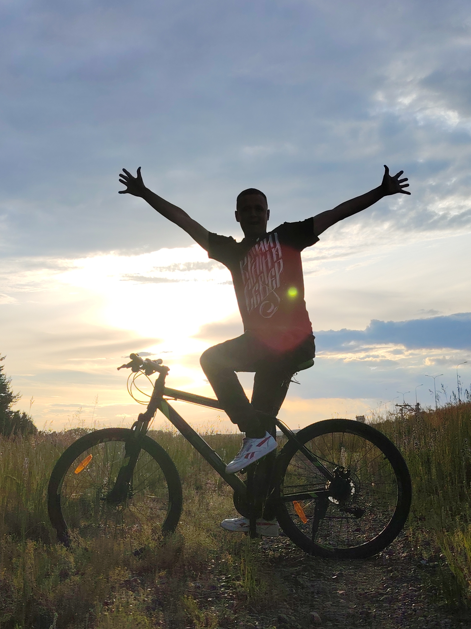 A silhouetted man sits on a bicycle with his arms outstretched, looking towards the sky. The sun is setting, casting a warm glow across the clouds and illuminating the scene from behind. The man is wearing a dark t-shirt with white text and dark pants, and white sneakers with red accents. The bicycle is dark green and appears to be a mountain bike. They are situated in a grassy field with tall grass and dirt paths. In the background, there are indications of urban infrastructure, such as utility poles and distant structures, suggesting the location is on the outskirts of a town. The overall mood is one of freedom and joy, accentuated by the expansive sky and the setting sun. The text on the man's shirt is illegible due to the silhouette. The time of day is dusk, with a partly cloudy sky. The location is likely Chaikovsky, Russia.