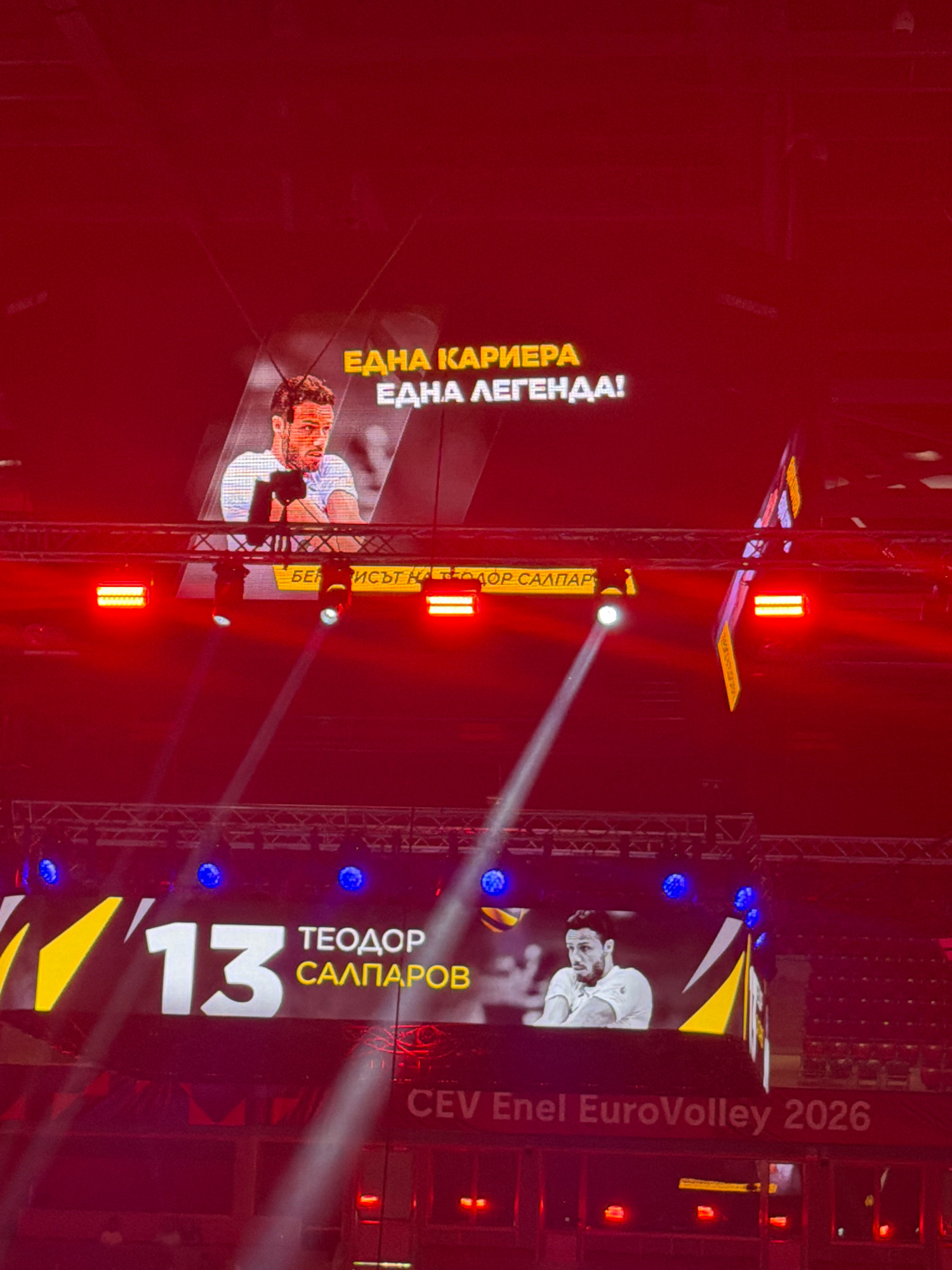 The image depicts a large screen display in a dimly lit indoor venue, likely an arena. The screen shows a portrait of a man, identified by the text "ТЕОДОР САЛПАРОВ" and the number "13" below him. Above the portrait, the text "ЕДНА КАРИЕРА ЕДНА ЛЕГЕНДА!" is visible in yellow and white lettering. Below this, a smaller yellow banner reads "БЕРИ ИСТ КА ТЕОДОР САЛПАР". At the bottom of the screen, the text "CEV Enel EuroVolley 2026" is displayed in white. The overall scene is dominated by red lighting and the glow from the screen. The presence of stage lighting trusses and blue spotlights suggests a sporting event or a performance. The context of "EuroVolley" and "Sofia, Bulgaria" suggests this is a volleyball event held in Sofia.