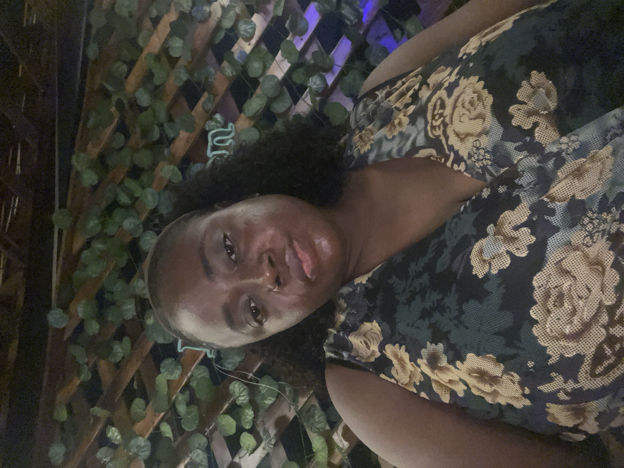 A selfie taken at night shows a Black woman with dark skin and curly hair posing in front of a wooden lattice wall adorned with artificial green leaves. She is wearing a dark floral print dress with light yellow and white flowers. In the background, a light blue neon sign is visible, and the overall lighting suggests an indoor or covered outdoor setting. The location is identified as Abuja, Nigeria.