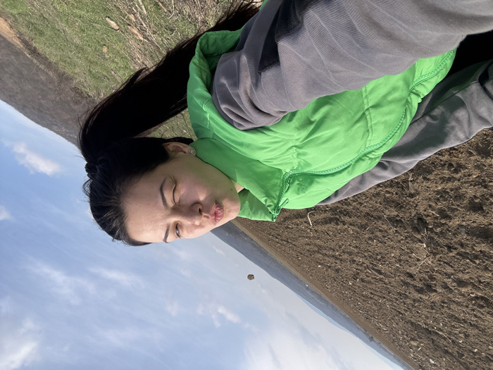 An angled photograph captures a young adult female engaged in a selfie, positioned against an outdoor rural landscape in the vicinity of Chubra, Bulgaria. The subject, situated in the lower left, has dark hair tied in a ponytail and is wearing a bright green quilted vest over a grey long-sleeved top. Her eyes are squinted, and her lips are puckered in a deliberate pose. The immediate foreground consists of dark brown, freshly tilled earth. Beyond this, a sloping grassy area leads to distant hills or mountains in the background. The sky overhead is blue with scattered white clouds. A small, light-colored, undefined object is visible in the upper right portion of the sky.