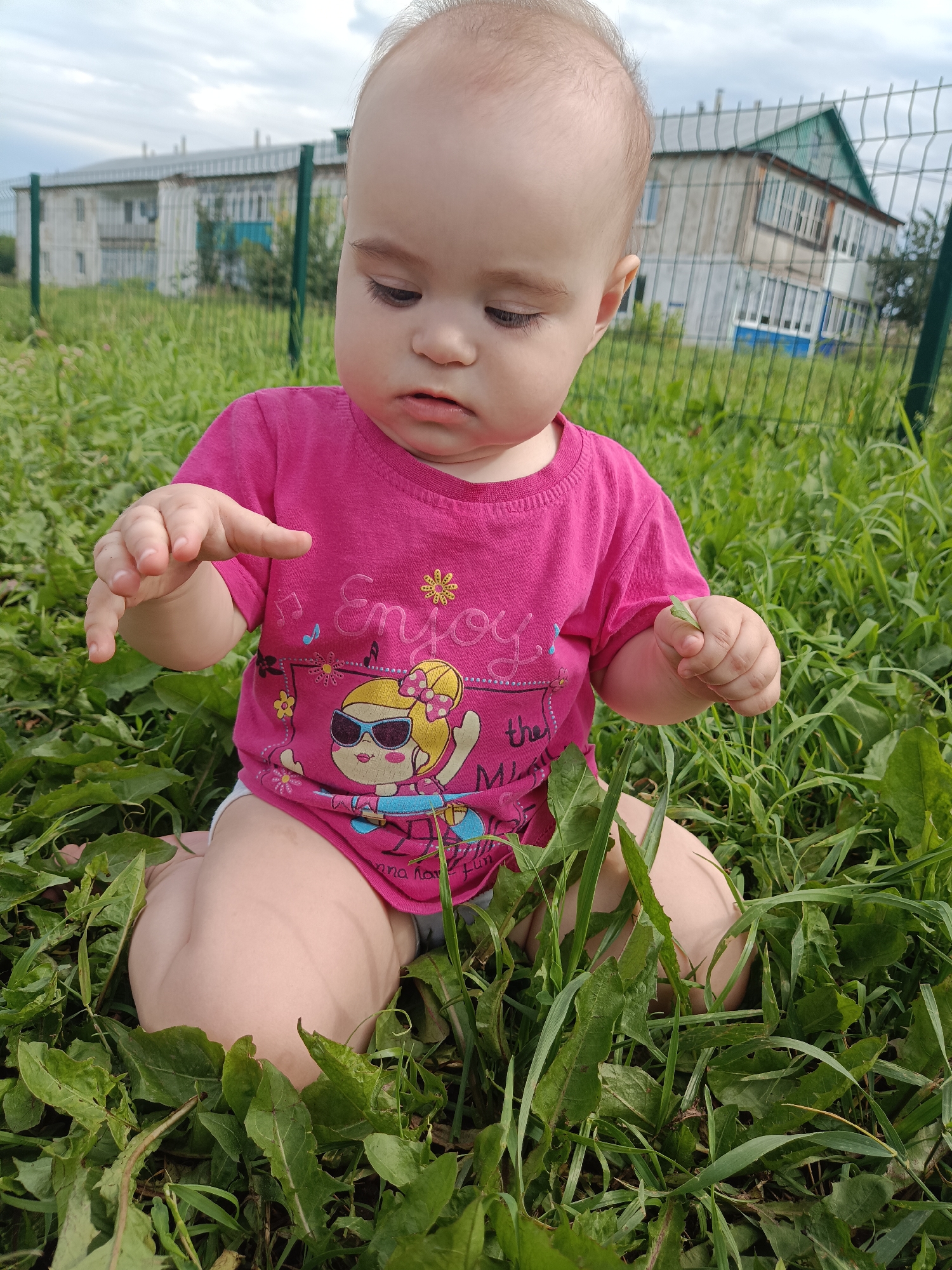 A young child, likely a toddler, is sitting in tall green grass. The child is wearing a bright pink t-shirt with a graphic print featuring a cartoon character and the word "Enjoy". The child's hands are visible, with one hand extended forward as if reaching for something, and the other holding a small green leaf. In the background, there is a metal fence and a two-story building with a green roof, indicative of a residential area, possibly in the city of Posad, Russia, given the context. The sky is overcast.
