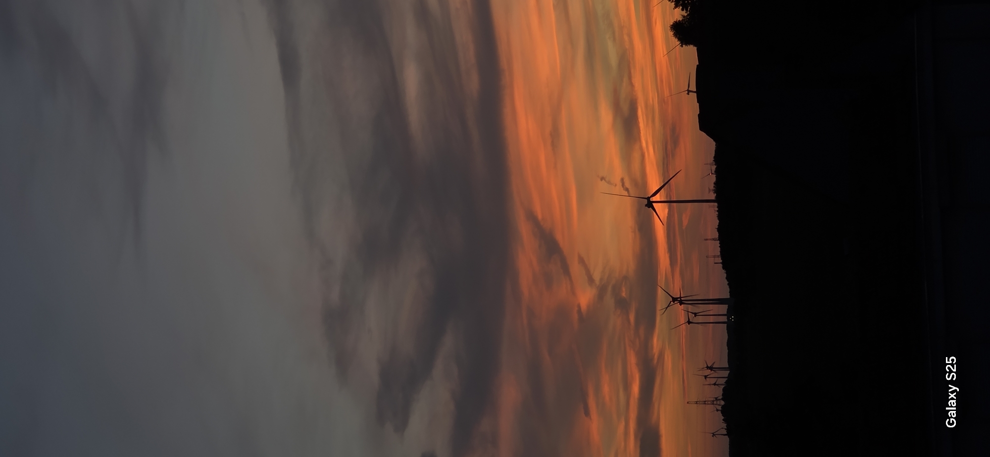 This is a vertical, full-frame image of a sunset over a landscape dotted with wind turbines and other structures, silhouetted against a dramatic sky. The sky transitions from a deep, dark grey at the top to vibrant orange and red hues closer to the horizon, with streaks of darker clouds intermingling with the brighter colors.
In the mid-ground, a hilly terrain is visible, entirely in silhouette. A multitude of wind turbines of varying sizes and designs are scattered across this silhouette. Some are tall and slender with distinct blades, while others appear shorter or are seen from different angles. In addition to the wind turbines, there are also what appear to be electricity pylons and some buildings or structures with angled roofs, also in silhouette.
The foreground is almost entirely black, obscuring any details of the ground. At the bottom left of the image, the text "Galaxy S25" is visible in white, indicating the device used to capture the photo. The overall impression is one of a serene, yet powerful natural scene juxtaposed with human-made technology. The location is Eeklo, Belgium.