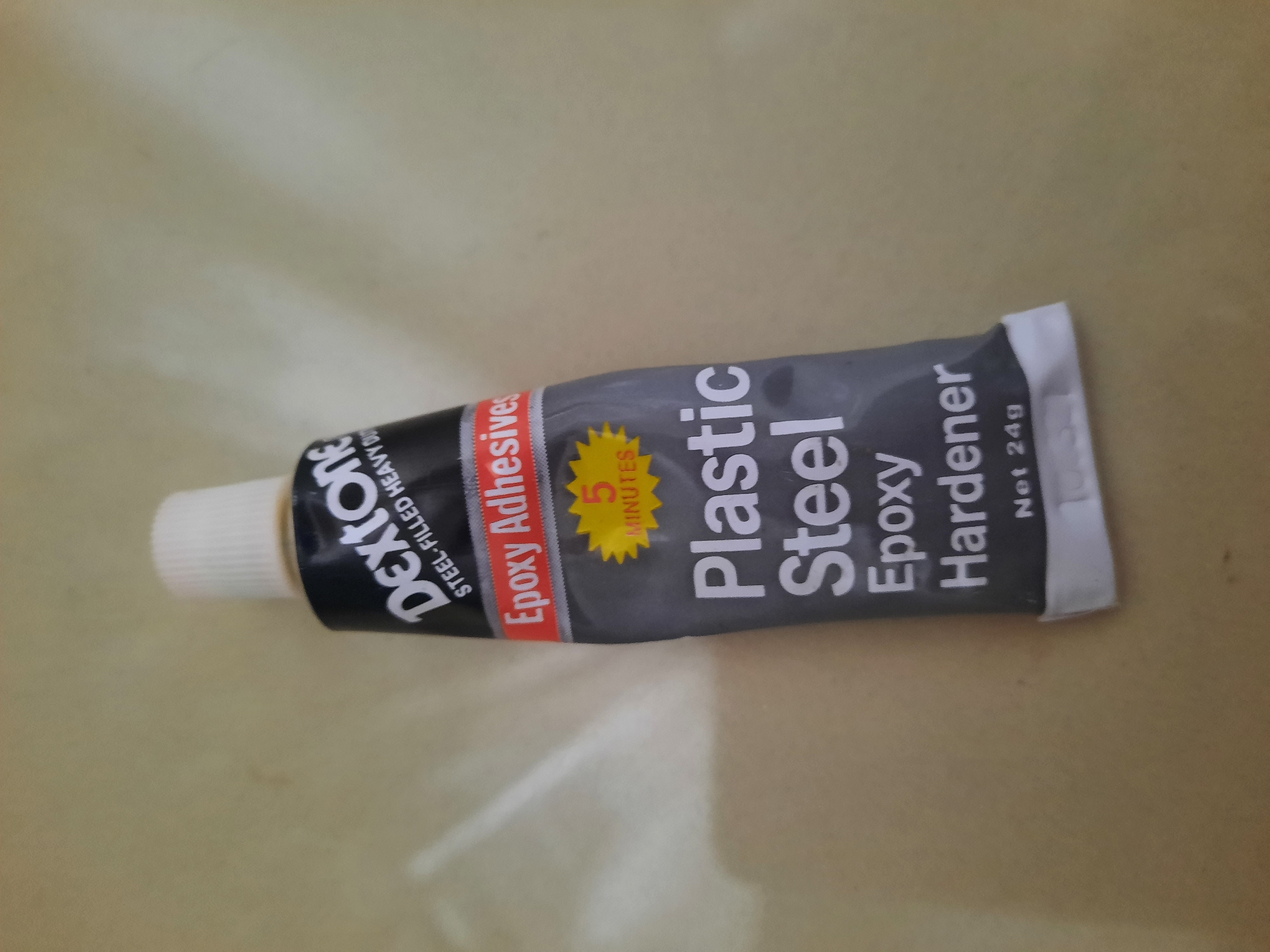 The image is a close-up, overhead shot of a tube of Dextone epoxy hardener. The tube is gray with black and white lettering. The brand name "Dextone" is prominently displayed at the top, followed by "Steel-Filled Heavy Duty". Below this, "Epoxy Adhesives" is written in red. A yellow starburst shape indicates "5 MINUTES". The main text on the tube reads "Plastic Steel Epoxy Hardener" in large white letters. At the bottom, "Net 24g" is visible. The tube appears to be placed on a light-colored, textured surface, possibly a table or countertop. No specific location in Lhokseumawe, Indonesia can be determined from the image.