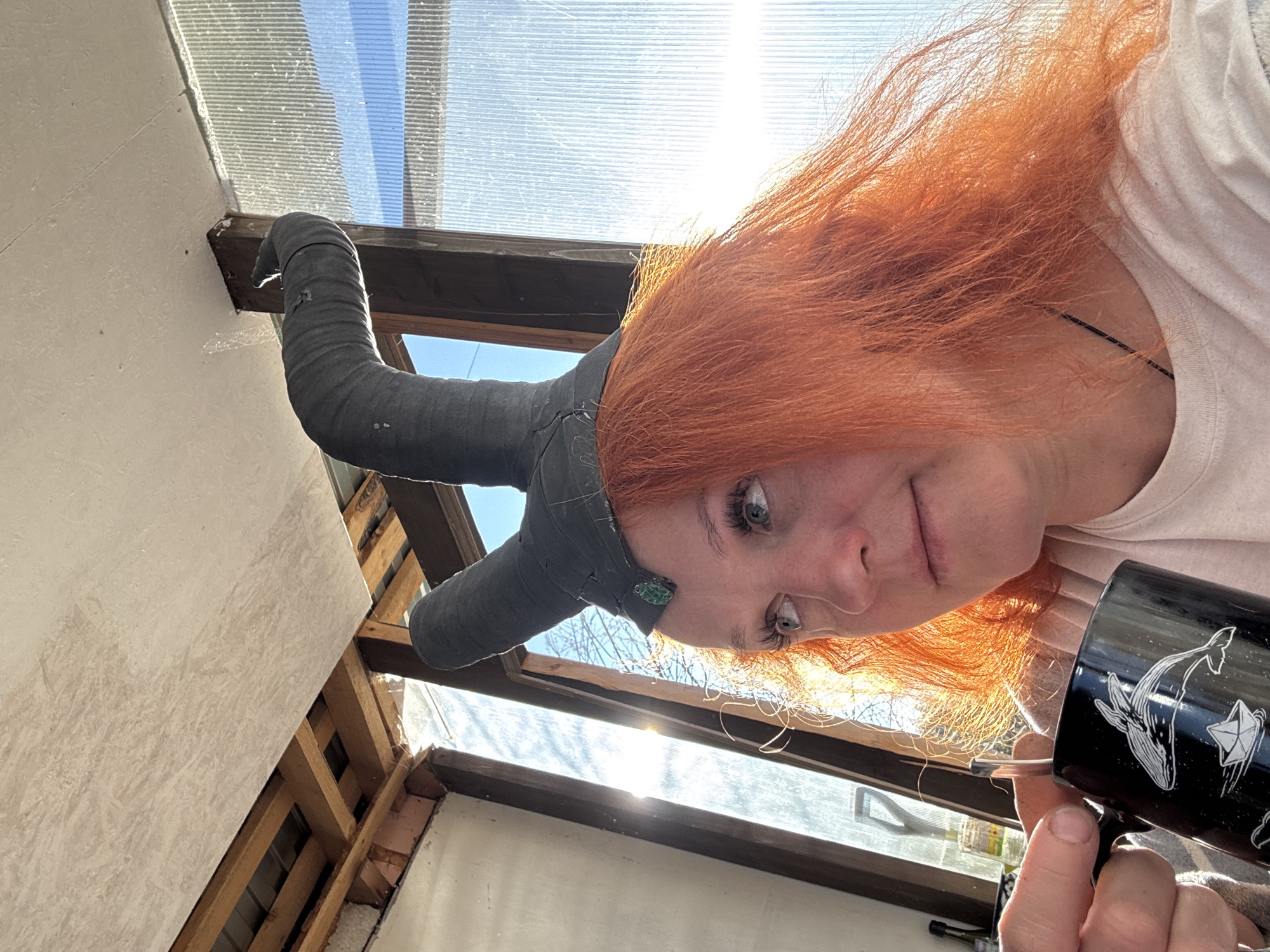A woman with red hair is holding a black mug with white whale and kite drawings. She is wearing a makeshift horned helmet. The background appears to be a partially constructed or renovated room with exposed wooden beams and a translucent roof. The location cannot be determined with available image information.