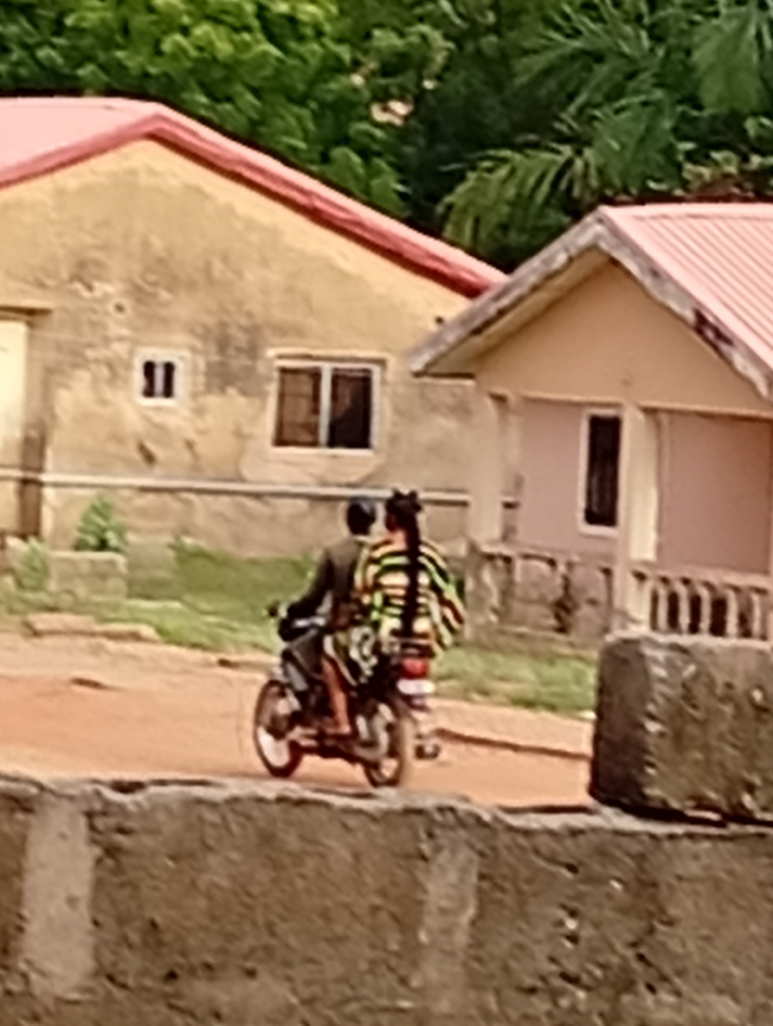 A daytime scene captures two people riding a motorcycle, commonly known as a boda-boda, on a dirt path in Adankolo, Nigeria. The rider is mostly obscured by the passenger, wearing a dark-colored top. The passenger, seated behind, is clearly visible from the back, showcasing long, dark braided hair, styled with two distinct sections at the crown. They are wearing a vibrant, striped garment featuring horizontal bands of green, yellow, and black.
In the background, two single-story houses with reddish-pink corrugated metal roofs are visible. The house on the left has a light, possibly cream or beige, exterior with two small rectangular windows. The house on the right is a lighter, peachy-pink color with one visible dark window and a short porch-like structure at its base. Dense green foliage and trees provide a natural backdrop behind and between the buildings. A rugged concrete or stone wall runs across the foreground, partially obscuring the lower section of the motorcycle and the path. The light suggests clear weather conditions. There is no visible text in the image.