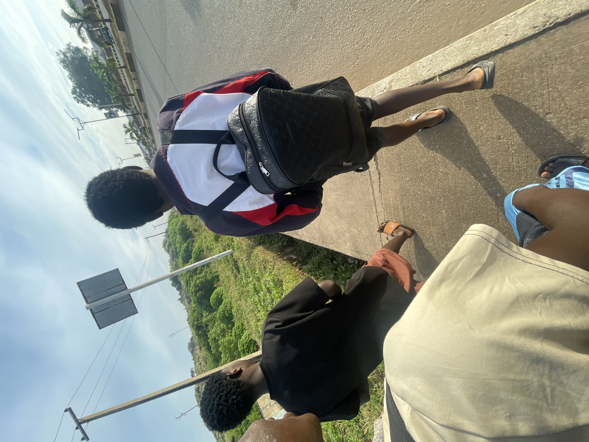Three individuals are walking along a paved sidewalk adjacent to a road. The individual in the foreground, partially visible, is wearing light-colored clothing. To their left, another individual is seen from the back, wearing a black t-shirt and orange sandals. In the center, a third individual walks ahead, also viewed from behind, wearing a red, white, and blue jacket and a black backpack with a patterned design. This individual is wearing dark shorts and black sandals. The sidewalk is made of concrete, and beyond it lies a road with some vegetation and trees on the left side. In the background, a clear sky with scattered clouds is visible. The scene appears to be in an urban or semi-urban environment, possibly in Jos, Nigeria, given the context. There are utility poles and streetlights along the road. The time of day appears to be morning or late afternoon due to the lighting and shadows.
