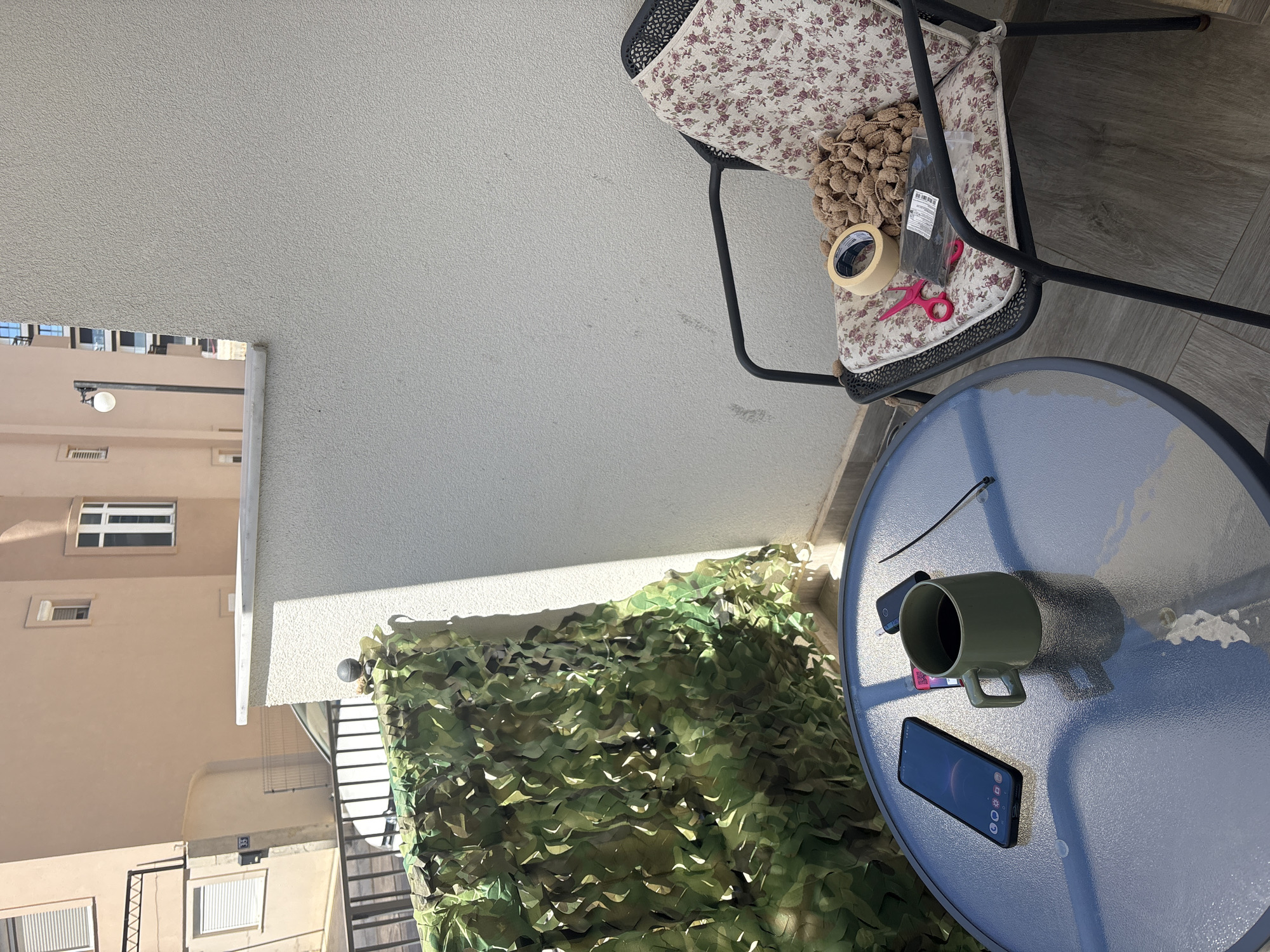 The image shows a balcony setting in Bar, Montenegro. A small metal table with a glass top holds a smartphone, a green ceramic mug containing a dark liquid, and a black zip tie. Next to the table is a chair with a floral cushion, on which are placed a bag of dried goods, a roll of tan tape, and a pair of pink scissors. Green camouflage netting is visible along the edge of the balcony, providing a visual barrier. In the background, the exterior of a beige multi-story building with windows and a satellite dish is seen. The setting indicates an outdoor living space with personal items in use.