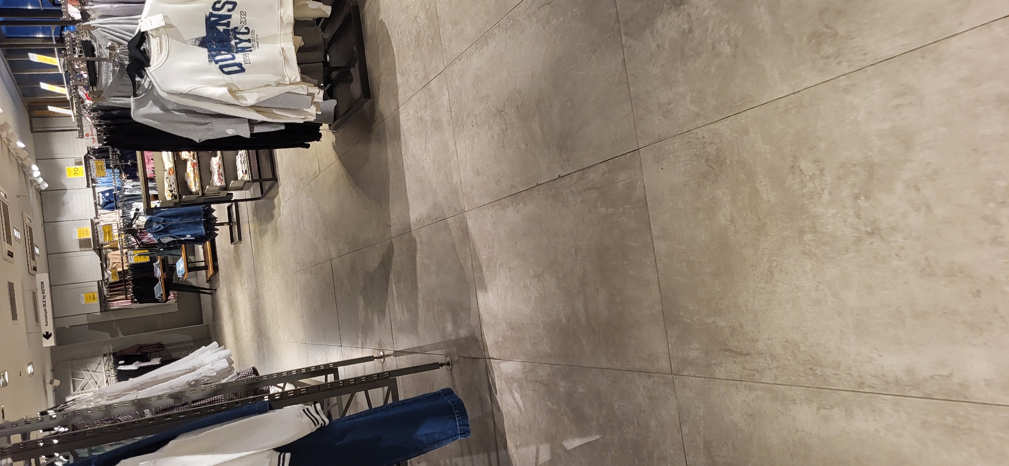 The image shows the interior of a clothing store, likely located in the city of Oskemen, Kazakhstan, as indicated by a sign reading "Koton OLE by KOTON". The store features racks of clothing, display shelves, and a tiled floor. The lighting is artificial, typical of an indoor retail environment. There are no people visible in the image. The clothing on display includes a variety of items such as sweaters, jeans, and shirts, with some items featuring price tags and promotional signs. One prominent item is a white sweatshirt with "QUEENS NYC" printed on it. The overall impression is of a well-stocked and organized retail space.