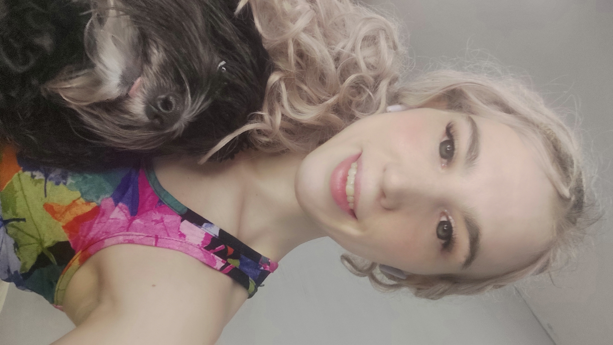 A pale-skinned woman with wavy blonde hair is smiling at the camera. A black and grey dog is lying on her left shoulder. The woman is wearing a colorful floral top. The background appears to be a plain white or light grey wall. Location information is not discernible.