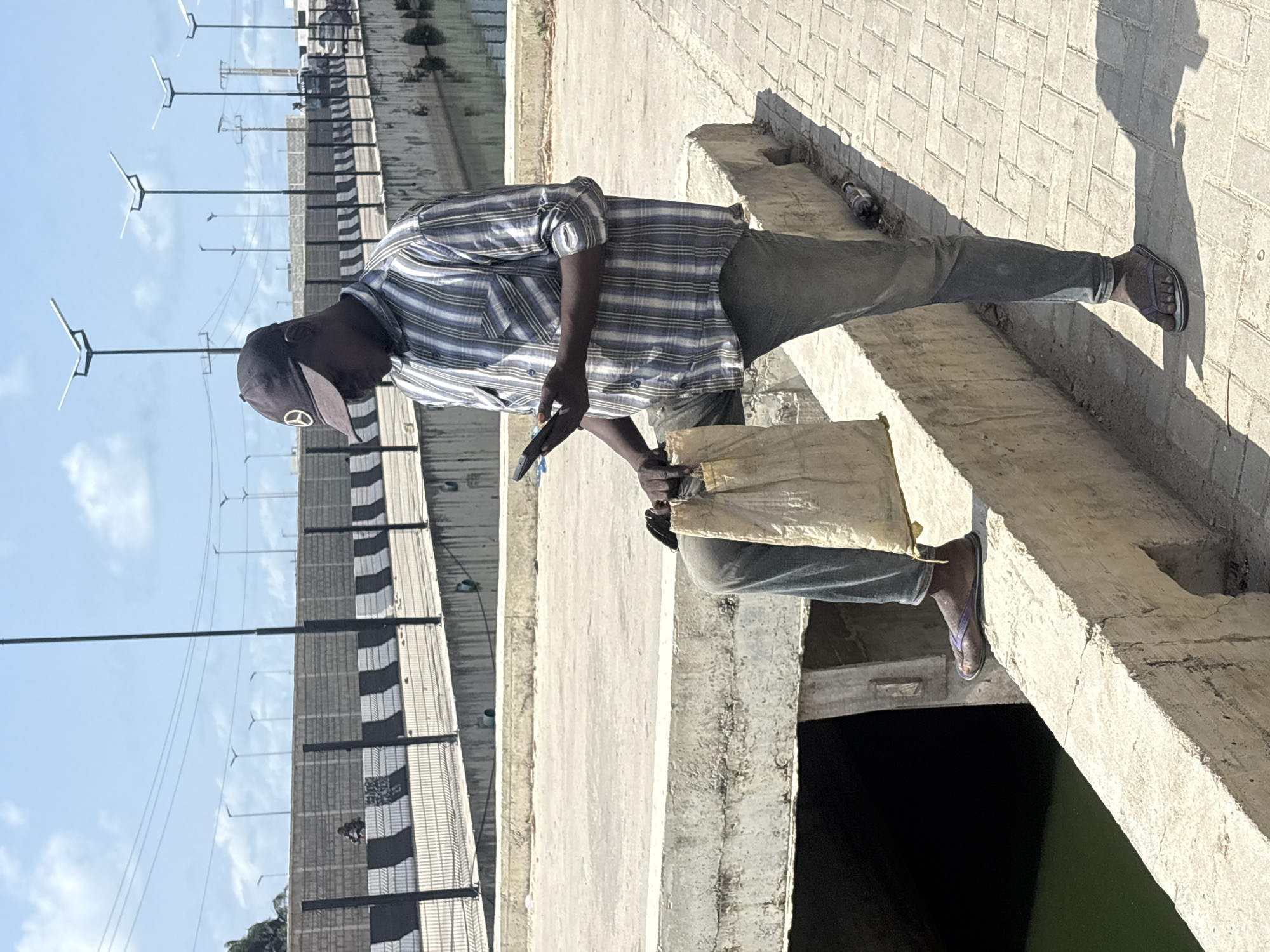 A man in a blue and white plaid shirt, gray pants, and a baseball cap stands on a concrete structure in Igboefon, Nigeria. He holds a cell phone in his left hand and a beige bag in his right. He is also wearing sandals. In the background, there is a fence, lampposts, and blue sky.