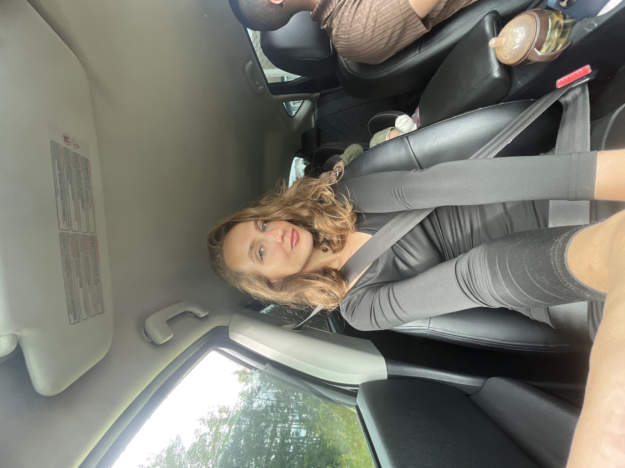 A woman is pictured inside a car in the city of Podshivalovo, Russia. She is sitting in the driver's seat, looking at the camera. The woman has wavy blonde hair, blue eyes, and is wearing a black long-sleeved shirt. She has a seatbelt on. There are two children in car seats in the back of the car. A man is visible in the passenger seat. The car appears to be moving, with trees and foliage visible through the windows.