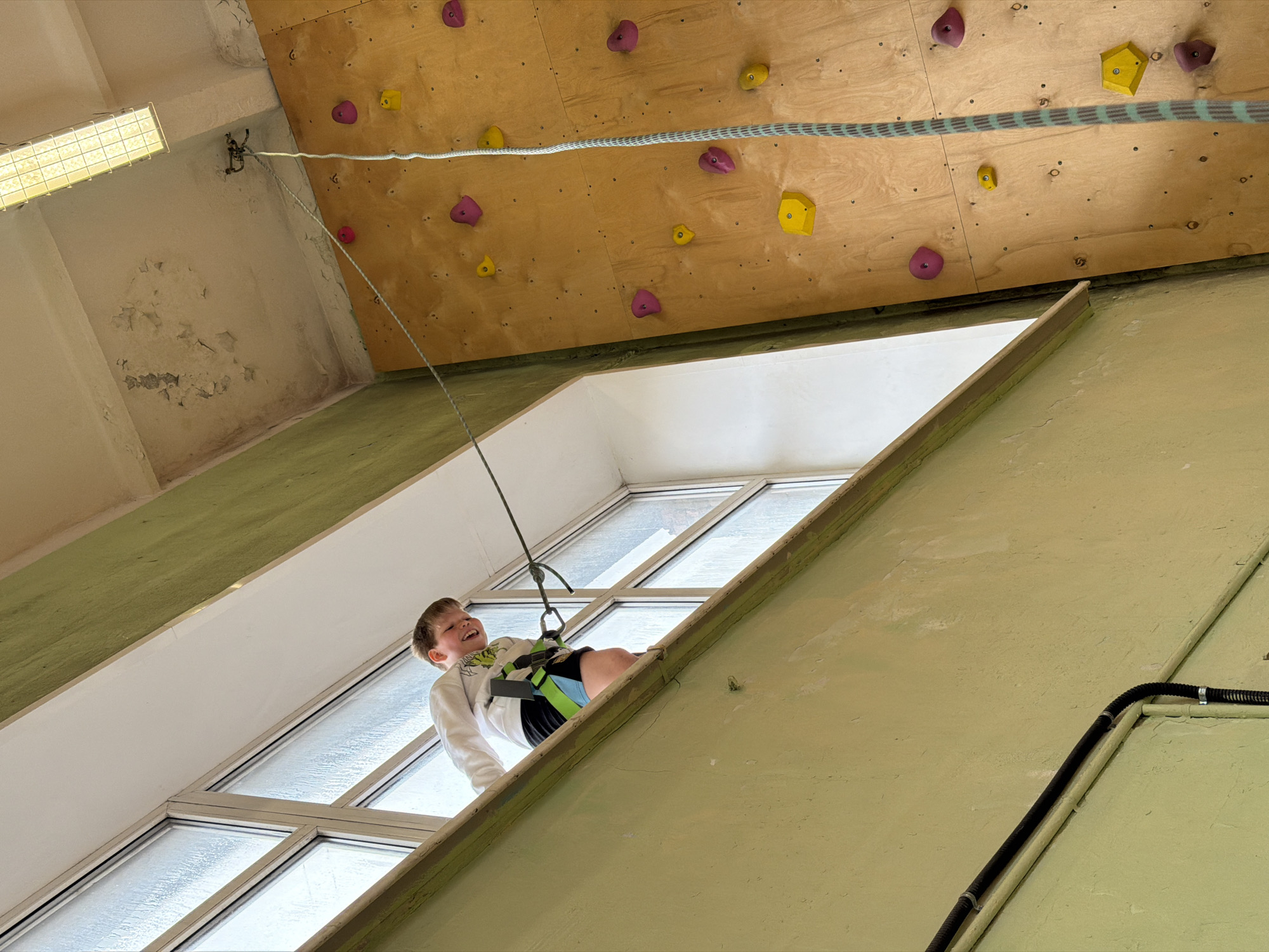 A low-angle indoor shot captures a young boy participating in a climbing activity, likely in Izhevsk, Russia. The boy, estimated to be between 6 and 10 years old, has short brown hair and is wearing a white long-sleeved shirt with a green design, black shorts, and a climbing harness with green and blue accents. He is attached to a grey climbing rope with light blue stripes, which extends upwards past a belay device or anchor point mounted high on the wall. The boy is smiling broadly, looking upwards with an expression of joy or excitement.
He is positioned in front of a large, multi-paned window section, which appears to be a skylight or high-mounted window, letting in natural daylight. The glass is textured or frosted, diffusing the light. Below him, to the right, is a light green painted wall with visible texture and some signs of wear, including peeling paint or plaster. Another section of wall, painted a similar light green, runs diagonally across the lower right of the frame, with a black corrugated conduit pipe attached.
Above the boy, a light brown wooden climbing wall is visible, covered with numerous colorful climbing holds in yellow and pink/purple hues. The climbing wall is angled steeply, suggesting a challenging climb. Further up and to the left, a portion of the ceiling shows more peeling paint and a rectangular fluorescent light fixture. The overall condition of the walls and ceiling suggests an older indoor climbing facility or gym. No visible text is present in the image.