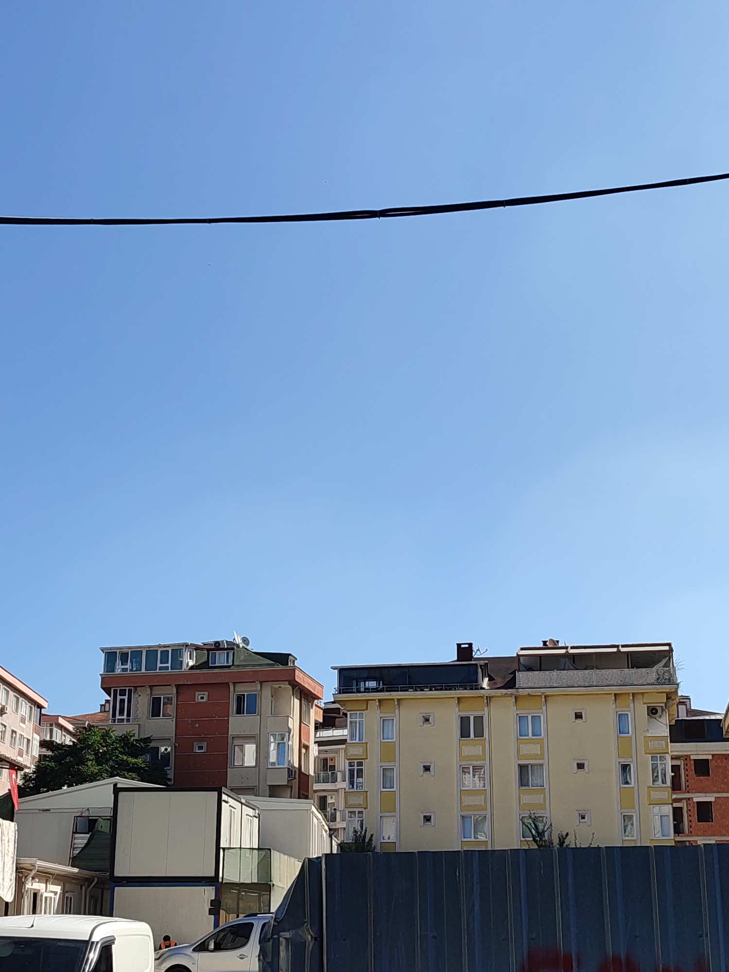 This image shows a street scene in Üsküdar, Turkey. In the foreground, a section of a dark metal fence is visible. Behind the fence, there are several portable buildings, some stacked on top of each other. To the left, a white van is partially visible. To the right of the portable buildings, a silver car is parked. In the background, there are several apartment buildings of varying heights and colors, including a tall red and orange building with many windows, and a yellow building with a large balcony area. The sky is clear and blue. There are power lines running across the top of the image. A small figure wearing an orange vest is visible near the silver car.