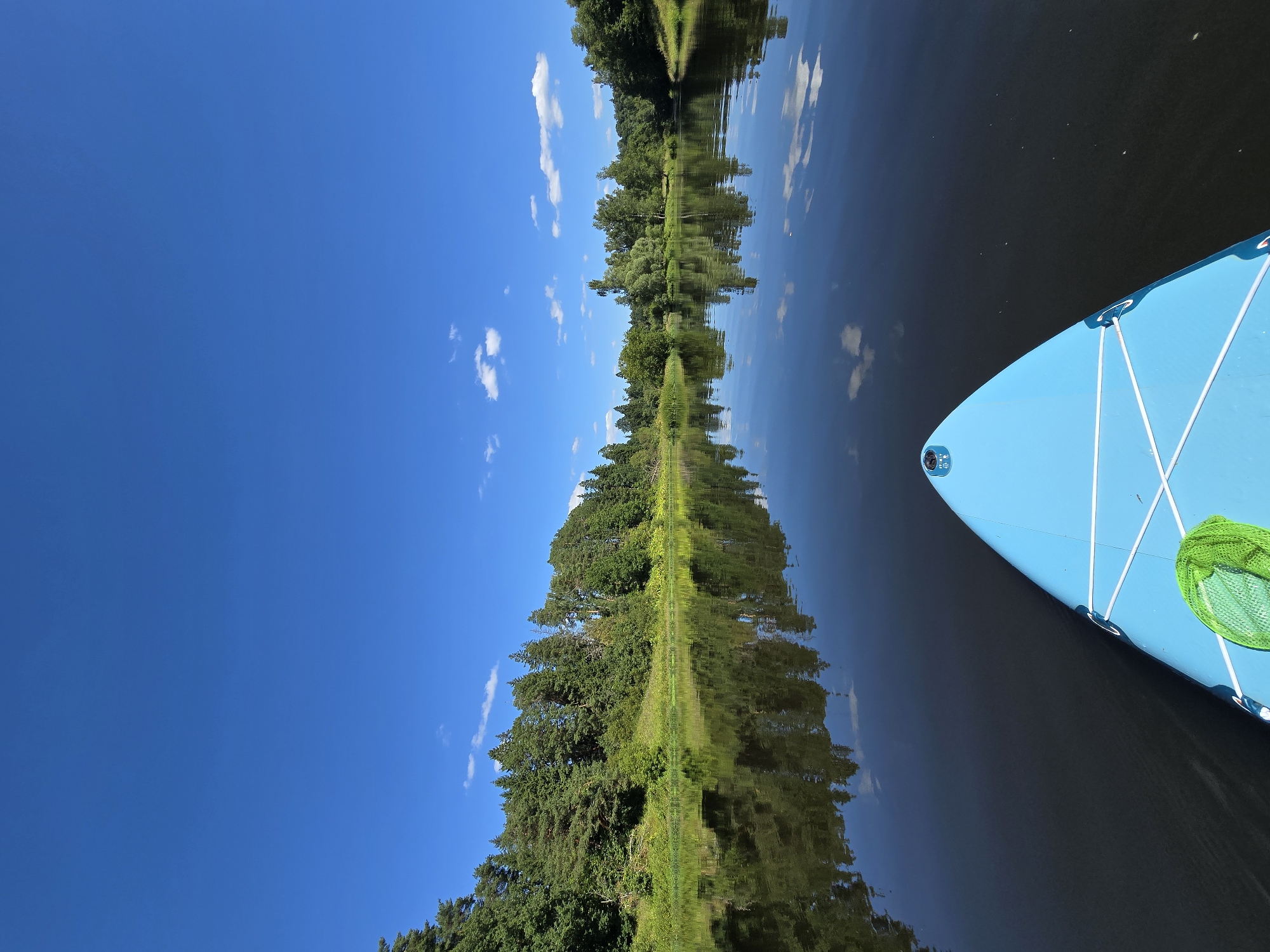 The image depicts a serene, wide river with lush green trees lining its banks. The sky is a clear, vibrant blue with a few scattered white clouds. The water is calm and reflective, mirroring the trees and sky. In the foreground, the nose of a bright blue stand-up paddleboard is visible, equipped with white bungee cords and a bright green net. The perspective is from the viewpoint of someone on the paddleboard, looking forward along the river. The location is the river in Voinskaya Chast' Mchs, Russia, a tranquil natural setting.