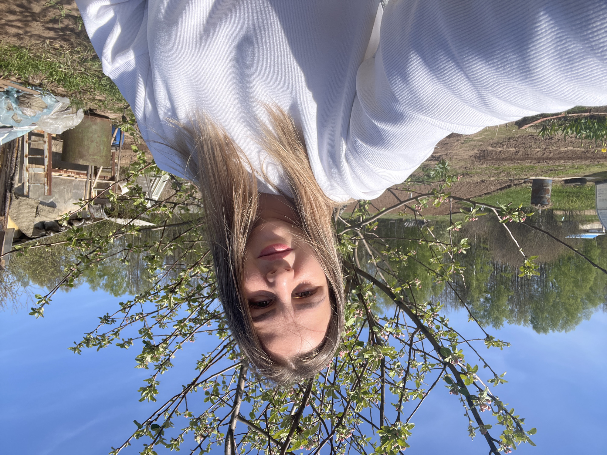 A woman is seen in an upside-down outdoor selfie. She is wearing a white sweatshirt. Her hair is blonde and shoulder-length. The background includes trees, a blue sky, and what appears to be farmland or a garden area. A building structure is visible in the background. The specific location within the city of Двигатель, Russia, cannot be determined from the visual information provided.