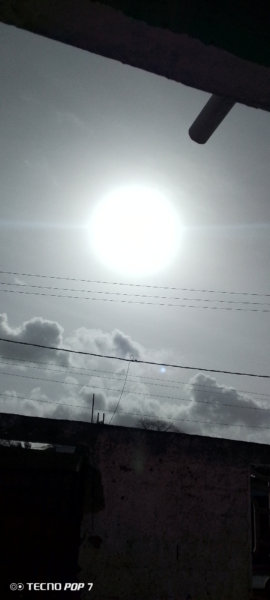 The image shows a black and white view of the sky in Altagracia, Venezuela. The sun is prominent and surrounded by a bright halo, indicating clear skies. Several layers of clouds are visible below the sun. Overhead power lines run across the middle of the frame. The lower portion of the image shows the edge of a building, with a partially visible window. A bare tree is situated on the roofline of the building. The image was taken using a TECNO POP 7 phone.