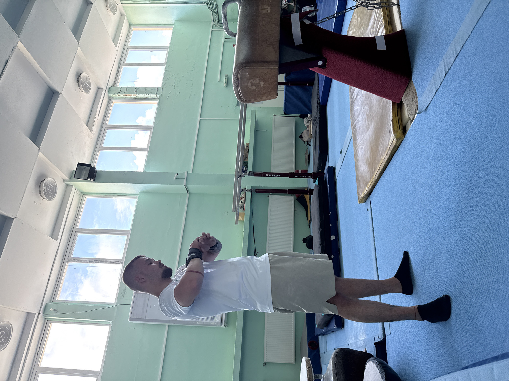 A man stands in a brightly lit gymnastics hall in Blagoevgrad, Bulgaria. He is wearing a white short-sleeved t-shirt, light beige shorts, and black socks. On his left wrist, he wears a black wrist wrap with the word "SKIDS" visible. His hands are clasped together in front of him, and he looks towards the right side of the frame, appearing to be observing or in a state of preparation.
The setting is an indoor gymnasium with light green walls and a light blue floor covered in sports mats. In the background, various gymnastics equipment can be seen, including a pommel horse with maroon padding and parts of parallel bars. The base of the parallel bars has text that reads "JANSSEN FR. TS". Radiators are mounted on the wall below large windows. The ceiling is white with a grid of square panels and circular ventilation units. Through the numerous high-set windows, a clear, sunny sky with some clouds is visible, indicating it is daytime.