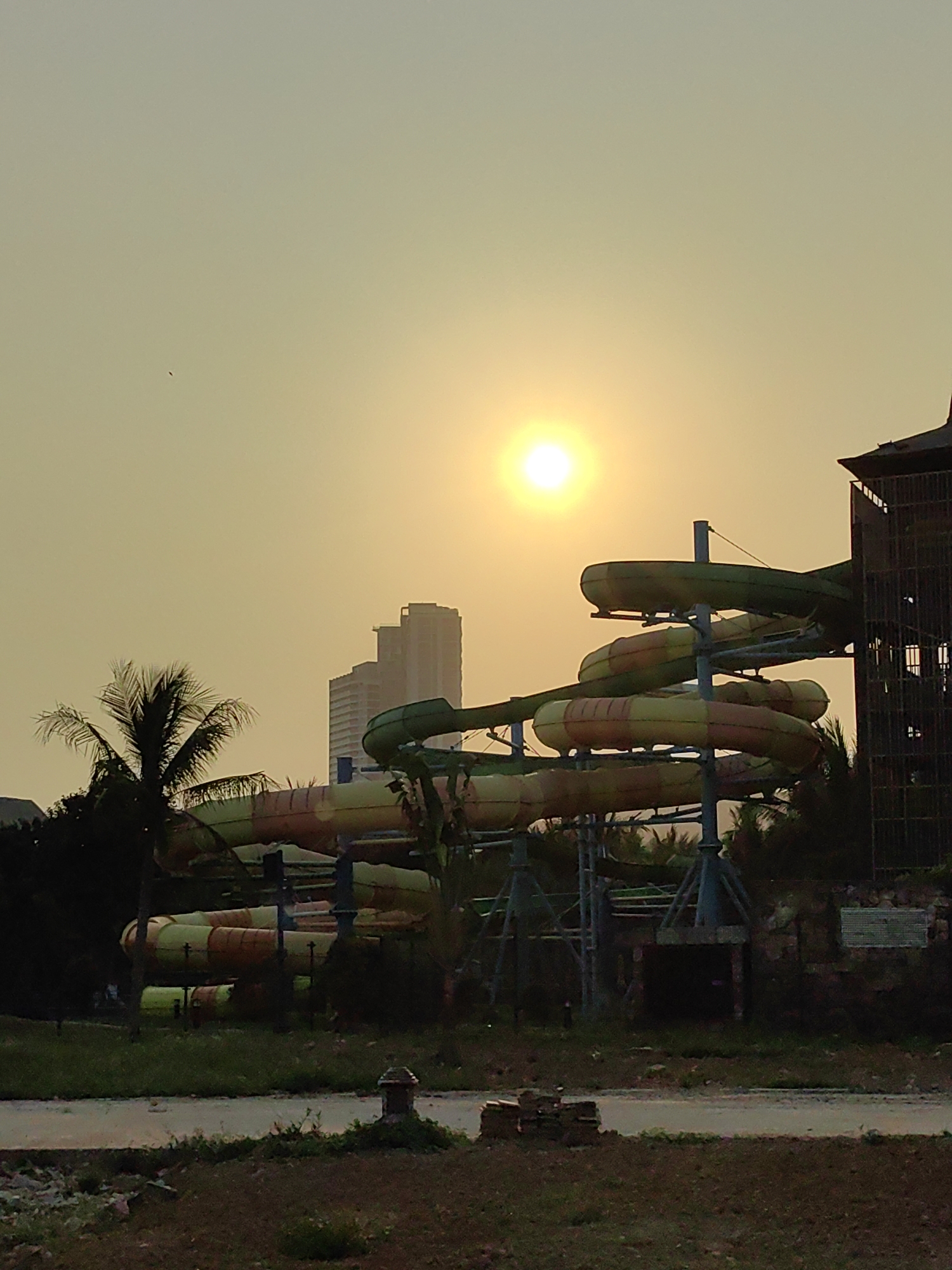 The image depicts a water park in Bai Chay Ward, Vietnam, at sunset. A multi-level water slide structure dominates the scene, with green, yellow, and orange tubes winding around support beams. Buildings are partially visible in the background, and a palm tree stands to the left. The ground in the foreground appears to be a mix of dirt and grass. The sun is positioned high in the sky, casting a golden glow over the landscape. A small bird is visible in the sky.
