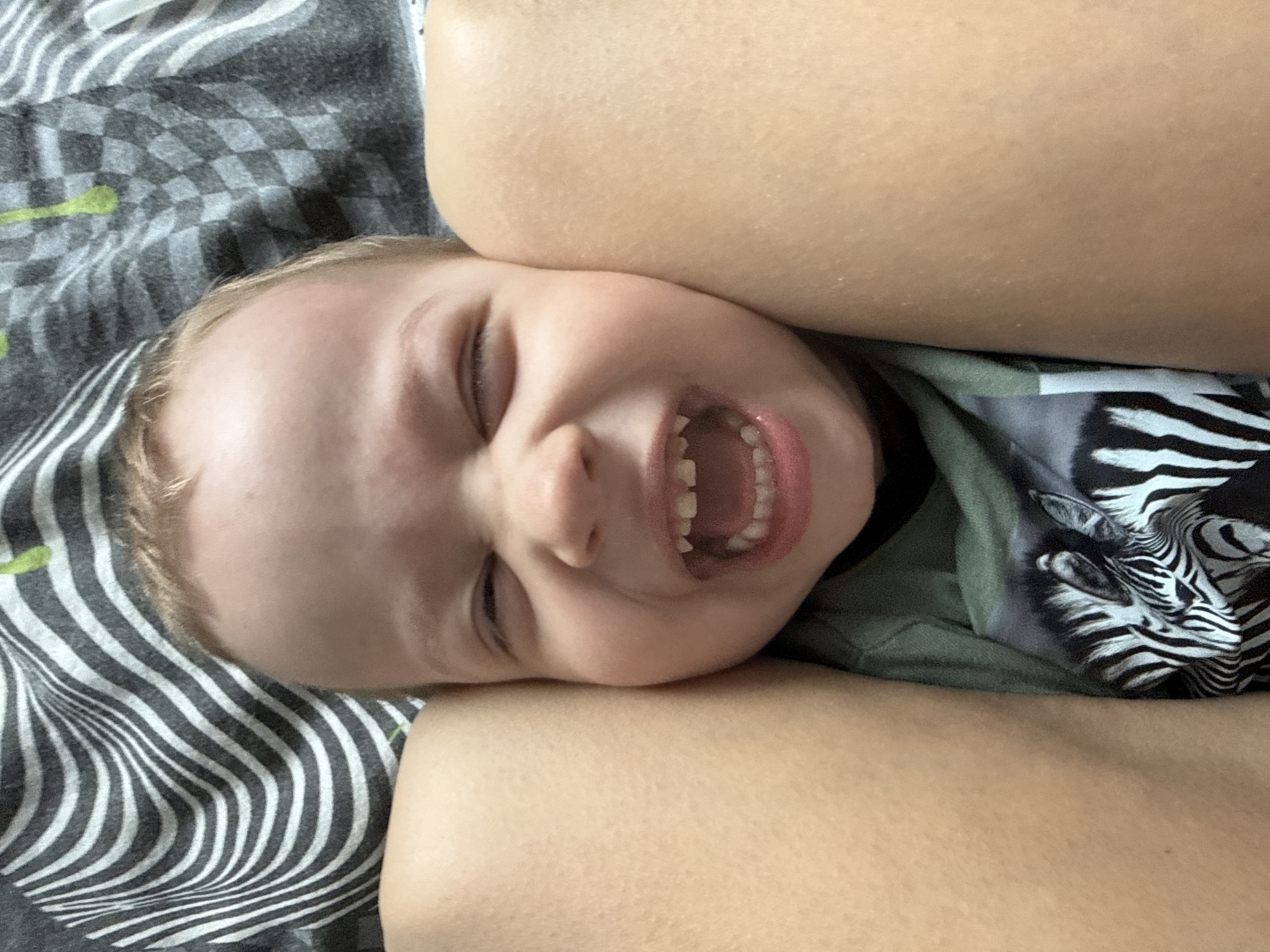 A close-up, high-angle shot captures a young child lying down, likely on a bed or soft surface, alongside an adult's body part. The child, with light-colored hair and fair skin, is positioned horizontally across the frame, with their head slightly turned to the right. Their eyes are squeezed tightly shut, and their mouth is wide open in a vivid expression of laughter or intense joy, revealing a full set of small, developing teeth. The child wears a green t-shirt that features a distinctive black and white zebra pattern on its right side.
The child's head rests against a patterned fabric, possibly bedding or a blanket, which displays a grey and white wavy stripe design on the left, partially adorned with faint lime-green leaf-like shapes. Further up and to the right, the fabric transitions to a grey and black checkerboard pattern. A section of an adult's bare arm or leg, with fair skin, is visible in the foreground, curving around the child's head, suggesting closeness or an embrace. No specific location cues, time of day, or visible text are present. The dominant emotion conveyed is one of intense happiness or playfulness from the child.