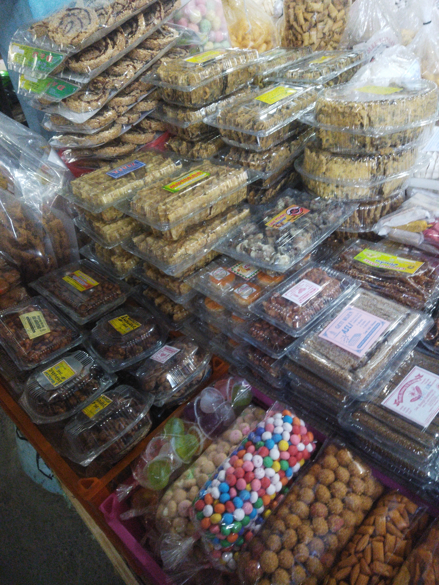 The image depicts a stall or market stall in Wonogiri, Indonesia, selling a variety of packaged snacks and sweets. The items are predominantly displayed in clear plastic packaging, stacked in rows and containers. Some packages contain what appear to be fried snacks, possibly including peanut brittle or similar confections, arranged in rectangular or circular shapes. Other packages hold smaller, crumbly items, and one prominent bag contains brightly colored round candies. Several packages feature labels with Indonesian text, indicating product names and possibly branding. The overall impression is one of a food vendor offering a diverse selection of treats.